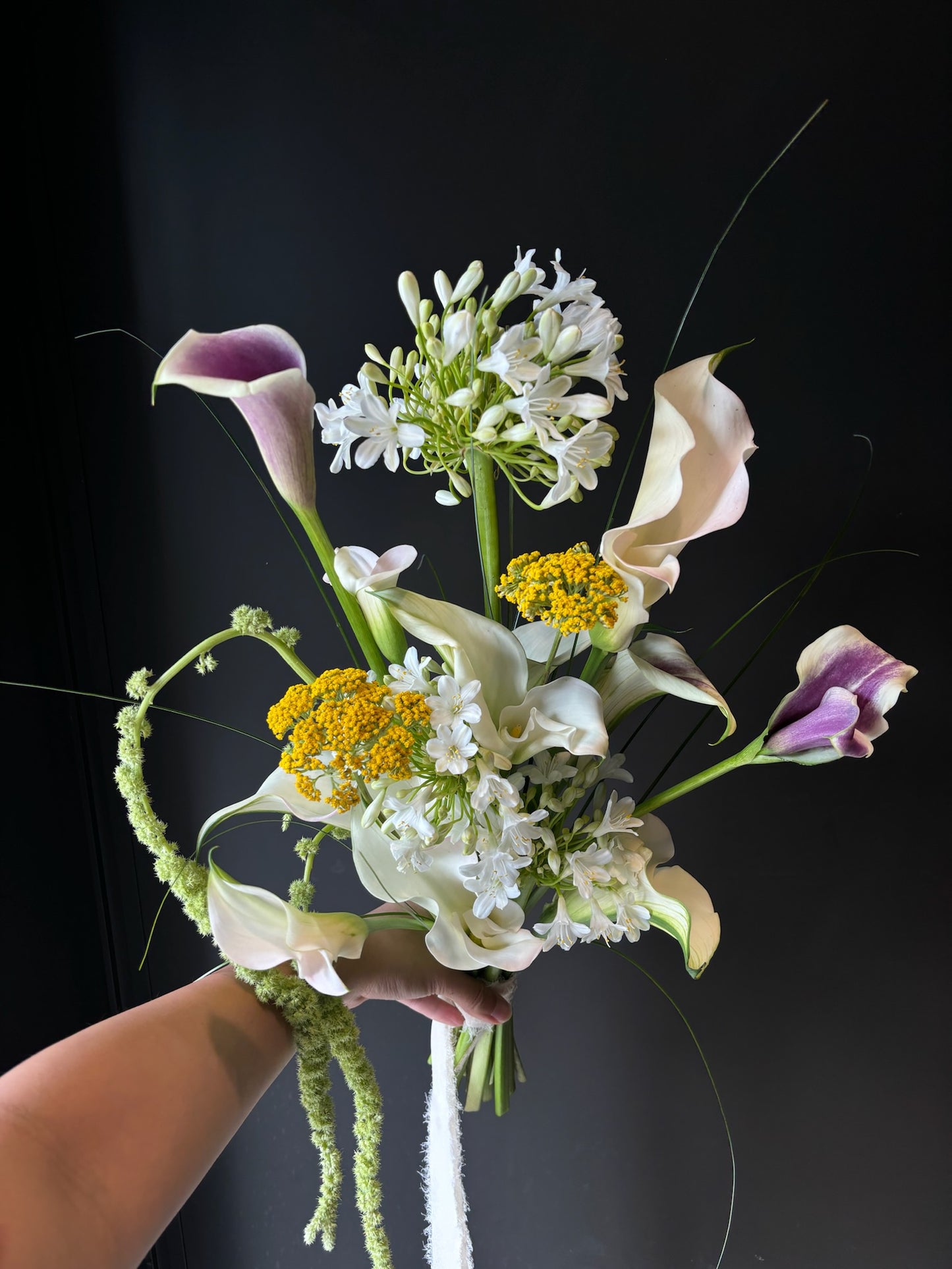 Hand holding a vibrant bouquet with white calla lilies, purple-edged calla lilies, yellow yarrow, and delicate white filler flowers against a dark background.