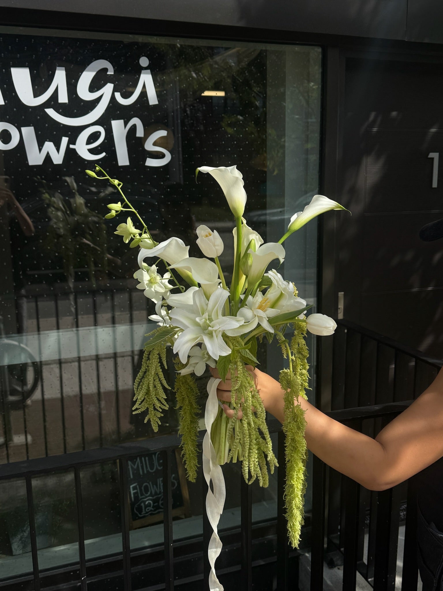 Hand holding a cascading white bridal bouquet with calla lilies, orchids, and trailing amaranthus in front of Miugi Flowers shop window