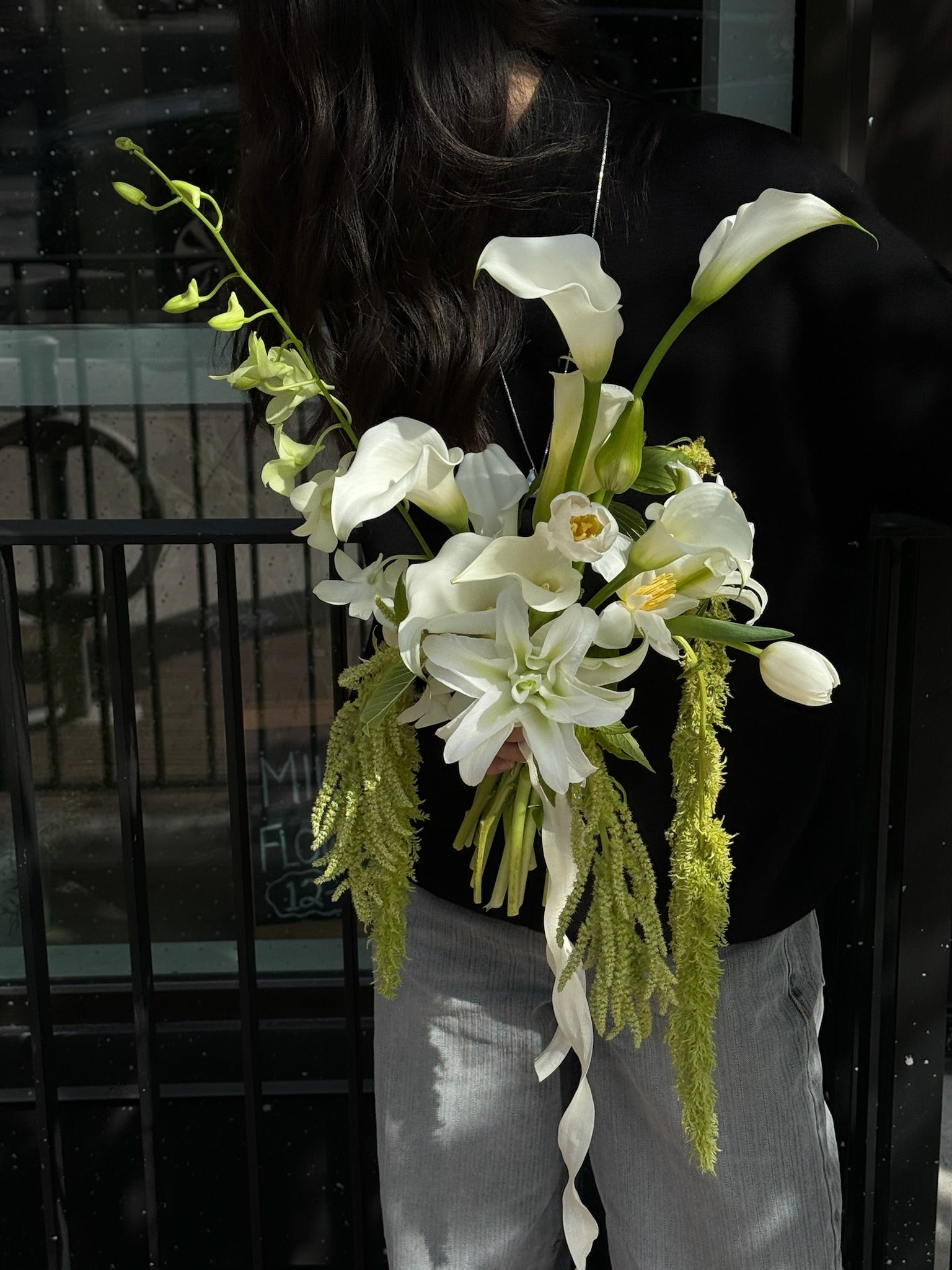 Person holding a cascading white bridal bouquet with calla lilies, orchids, and trailing green amaranthus, standing in front of a black background.