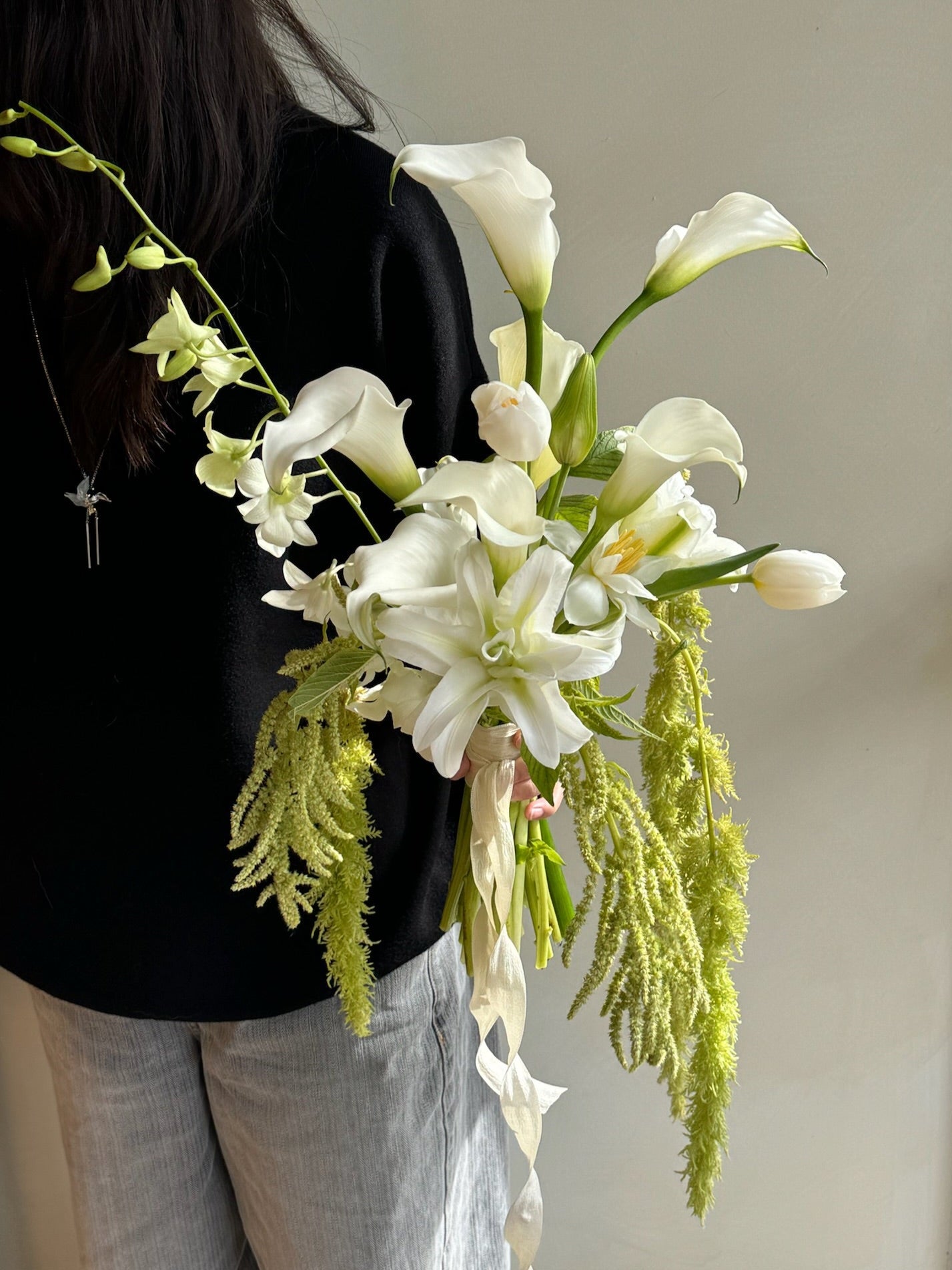 Person holding a white cascading bridal bouquet with calla lilies, orchids, and lush green amranthus against a neutral background