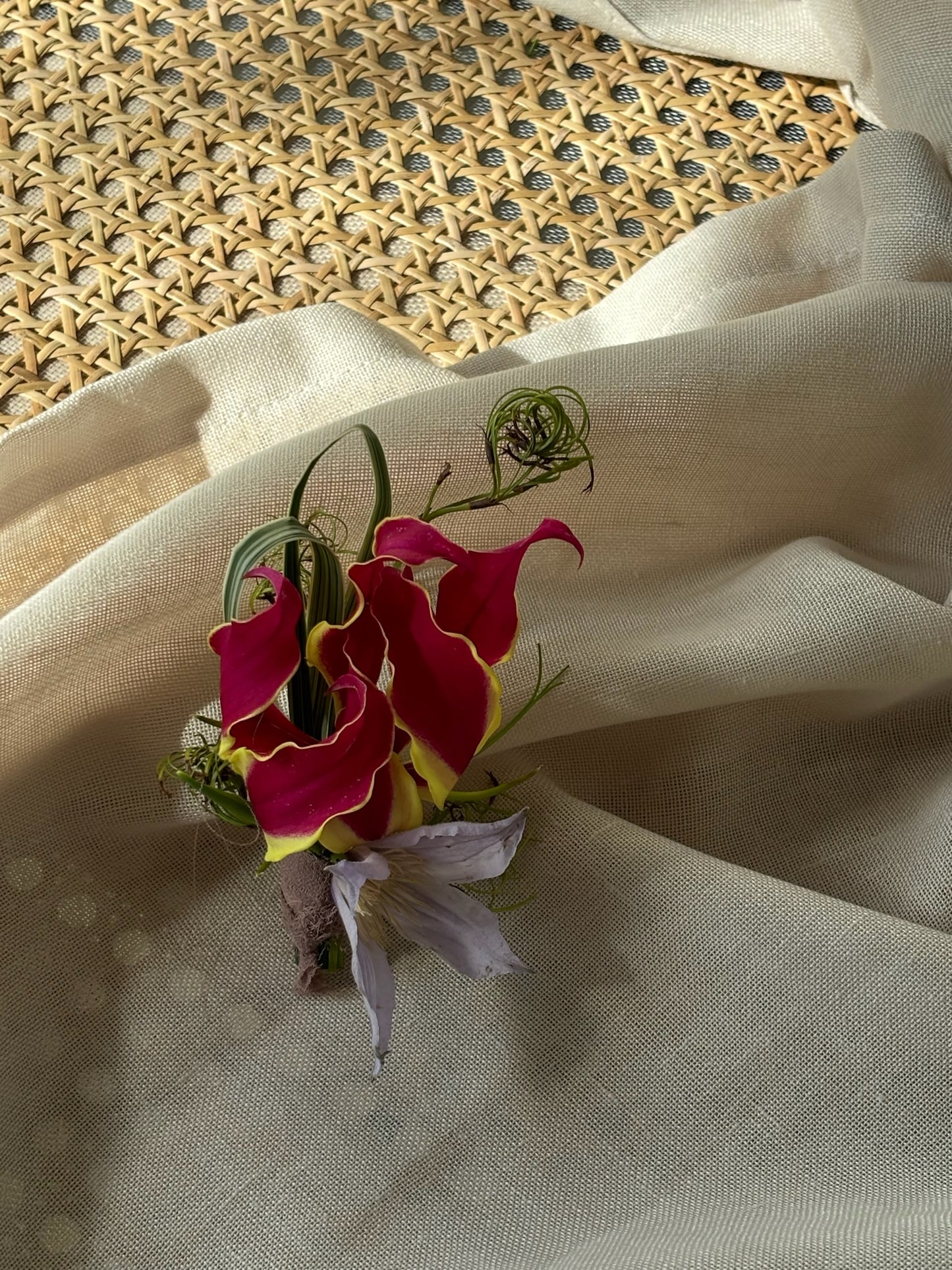 A striking boutonniere with deep red gloriosa lilies accented by green vine tendrils, resting on a soft beige fabric backdrop for an elegant presentation.