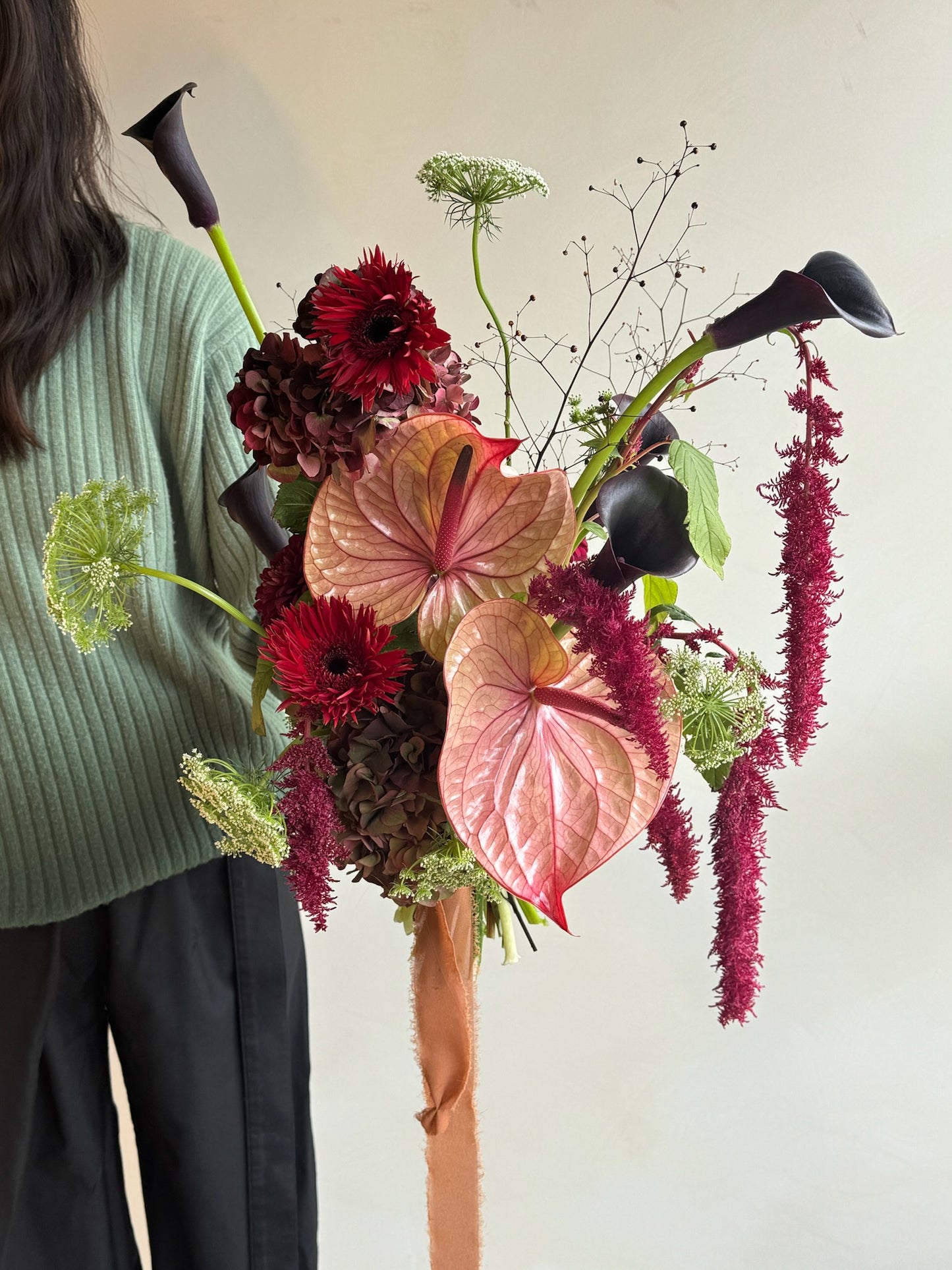 A large bridal bouquet with a long ribbon featuring pink anthuriums, dark red gerbera daisies, burgundy amaranthus, and black calla lilies, complemented by greenery and delicate filler flowers. The bouquet is held by a person in a green ribbed sweater and black pants, with a neutral indoor background.