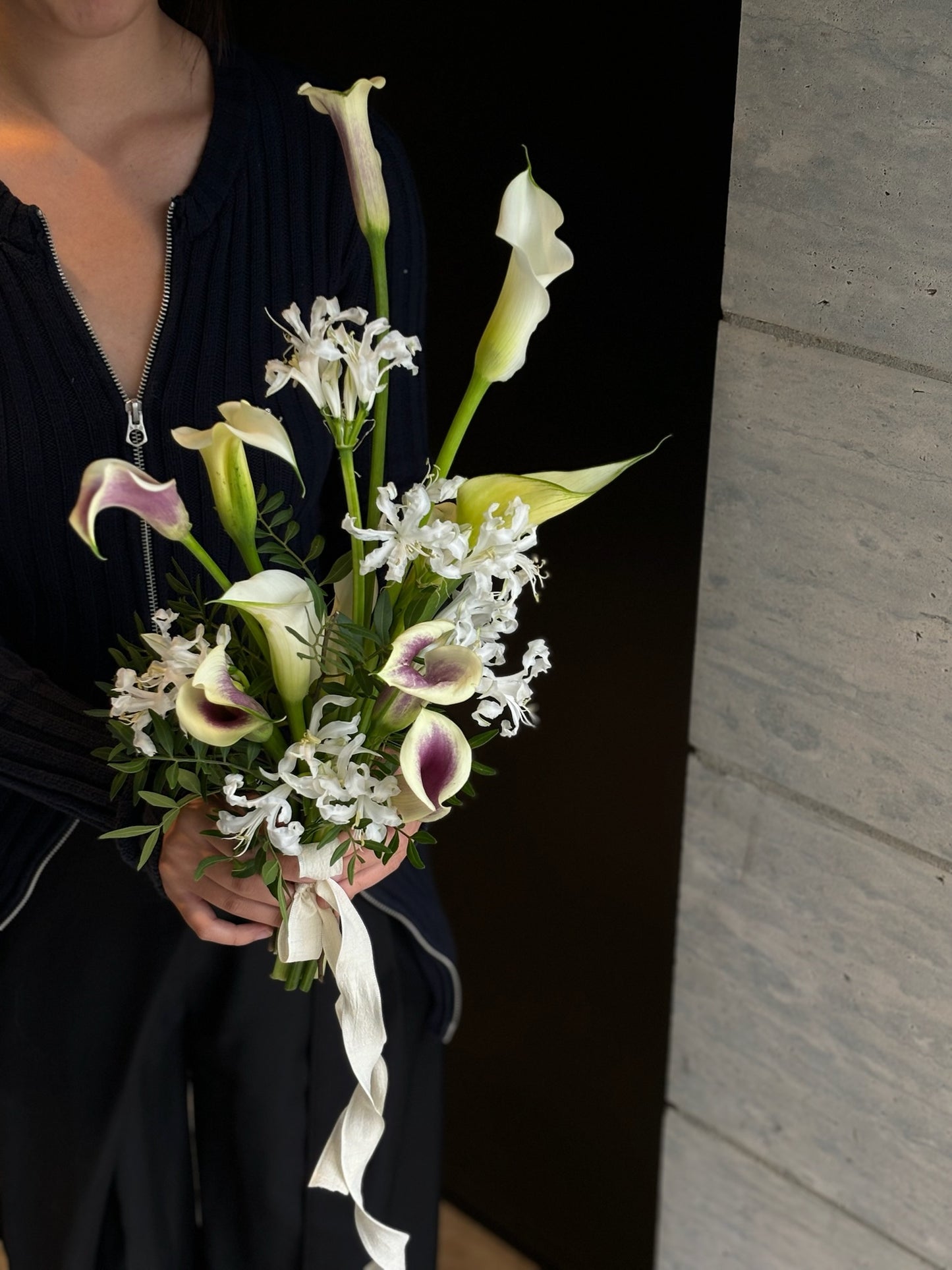 A person holding a bouquet featuring white calla lilies with purple edges, white dendrobium orchids, and white nerine flowers. The bouquet is accented with greenery and tied with a white ribbon. The person is wearing a dark outfit, and the background includes a light stone wall.