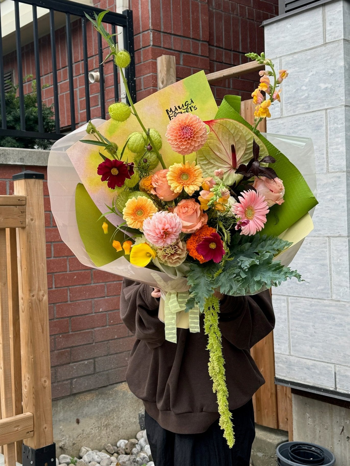 A large bouquet featuring pink gerbera daisies, peach garden roses, yellow ranunculus, green amaranthus, peach carnations, green ball dianthus, white anthurium, and deep burgundy dahlias. Wrapped in bright lime green and white paper, the arrangement is held outdoors in front of a brick wall and wooden fence.
