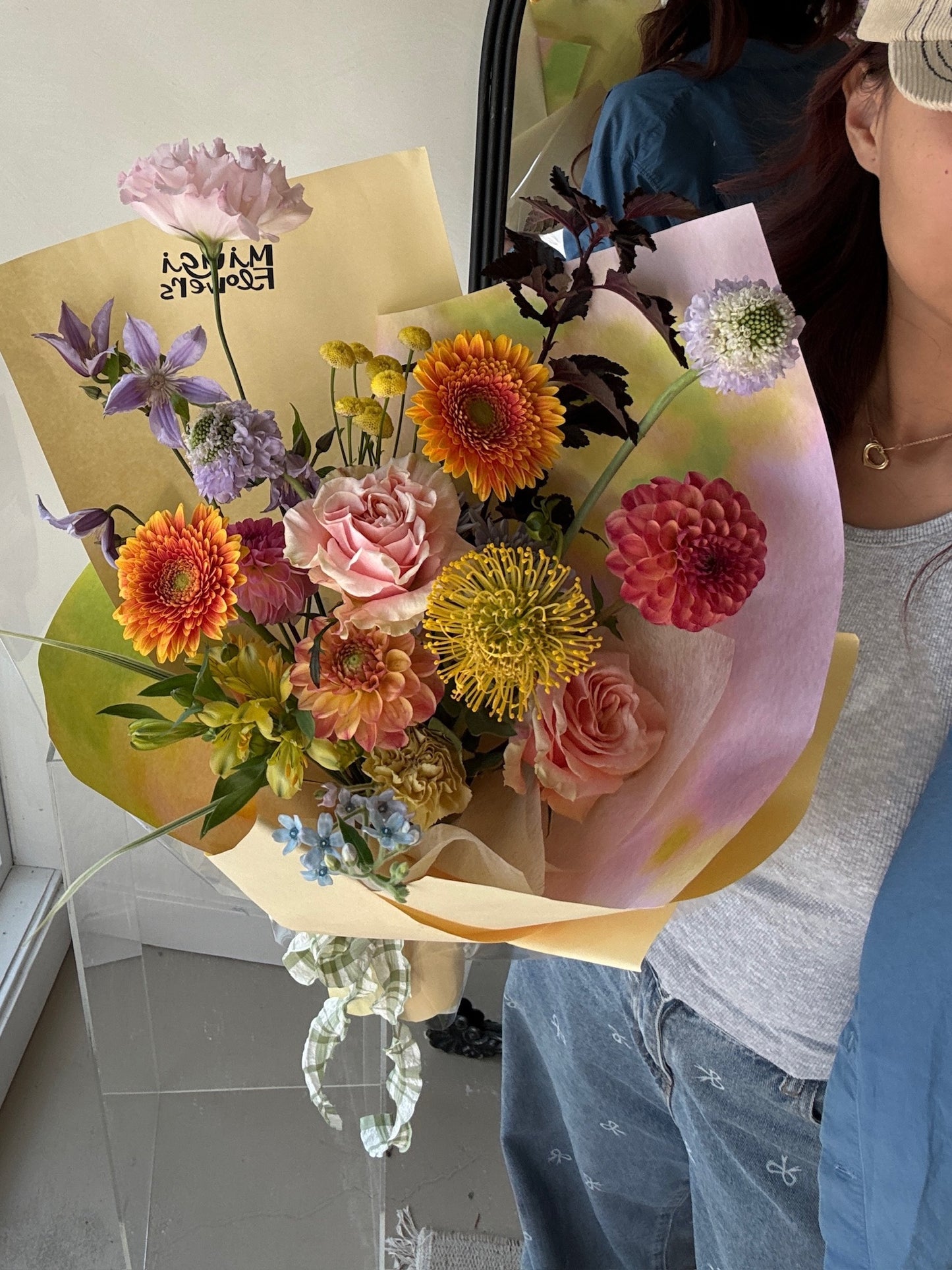 A bouquet featuring a mix of orange zinnias, yellow button chrysanthemums, pink spray roses, purple clematis, and green amaranthus, wrapped in light yellow and pink ombre paper. The arrangement is held against a mirror reflecting part of the bouquet and the person holding it.