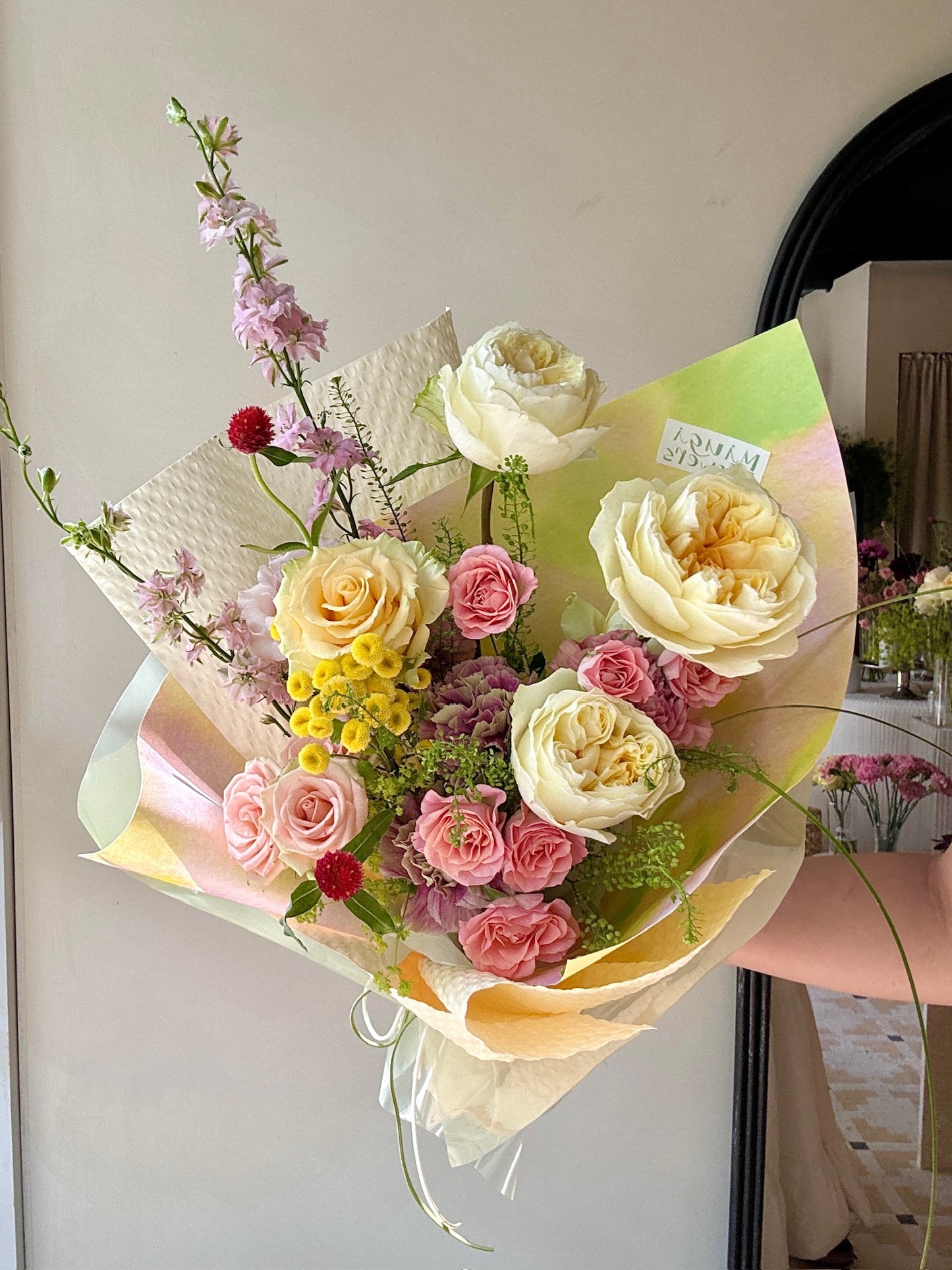 A bouquet featuring cream garden roses, pink spray roses, yellow button chrysanthemums, and red globe amaranth, accented with light pink astilbe and wrapped in pale green and beige paper. The bouquet is held in front of a mirror, reflecting a soft, natural light.