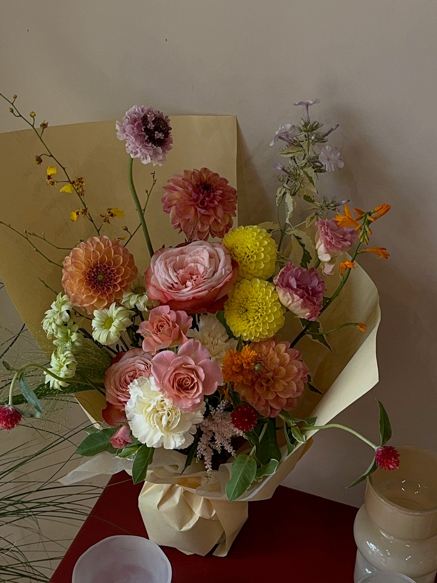 A bouquet with peach and pink garden roses, yellow button chrysanthemums, pink dahlias, and lavender delphiniums. The arrangement is wrapped in light yellow paper and placed on a red surface against a neutral background.
