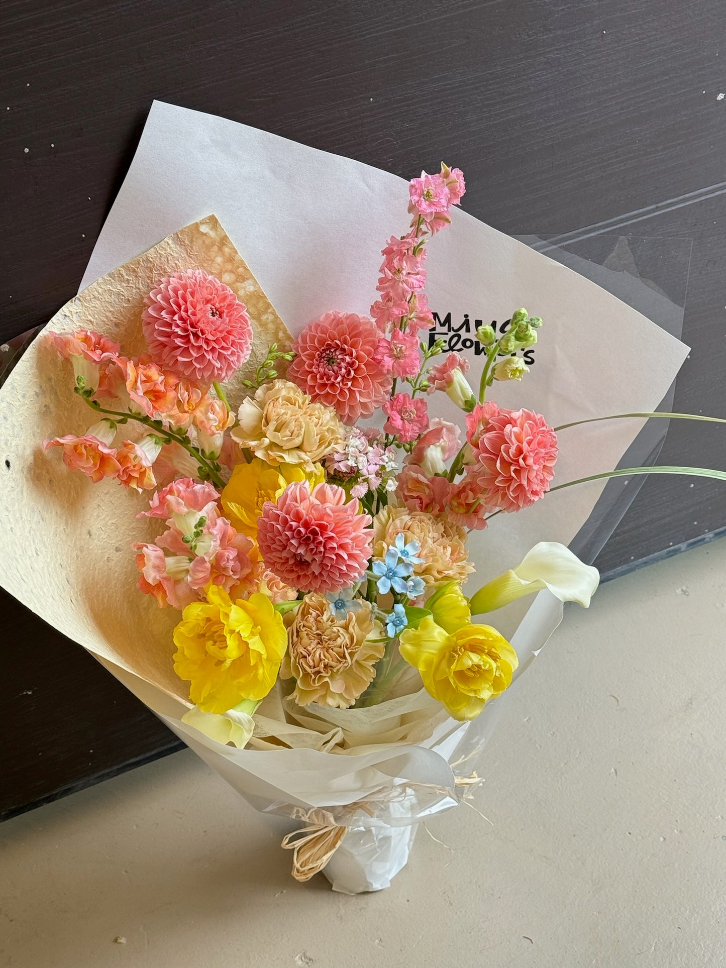 A bouquet featuring pink dahlias, yellow roses, carnations, and delicate pastel blue flowers. The arrangement is wrapped in light beige and white paper, set on a neutral-colored background indoors.
