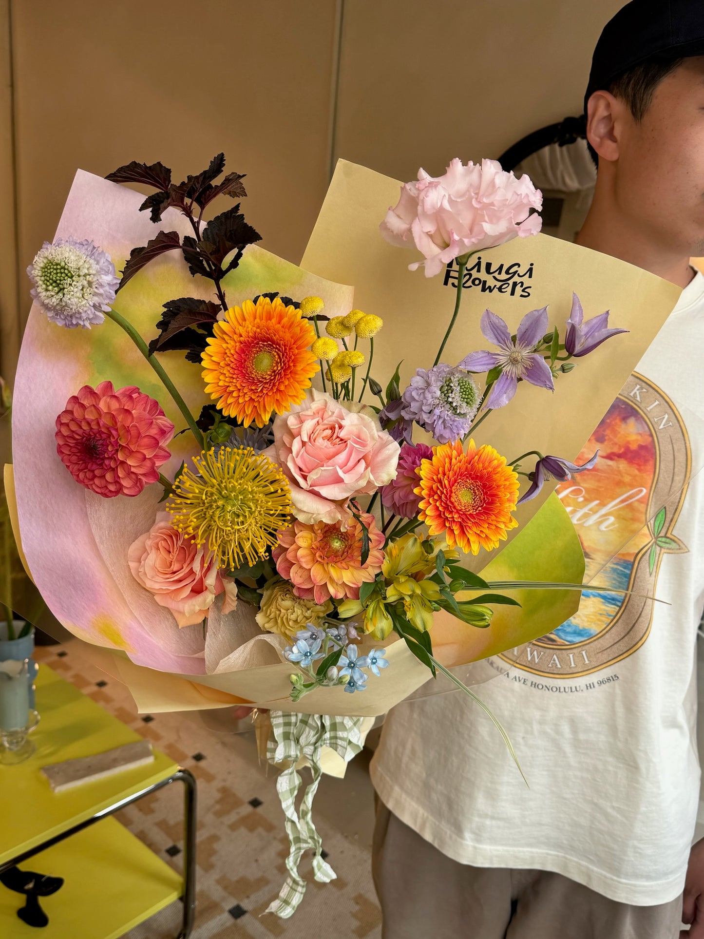 Bouquet featuring orange gerbera daisies, pink roses, purple clematis, yellow chrysanthemums, red dahlias, and green button mums. The arrangement is wrapped in yellow and pink paper, held by a person indoors next to a yellow table.