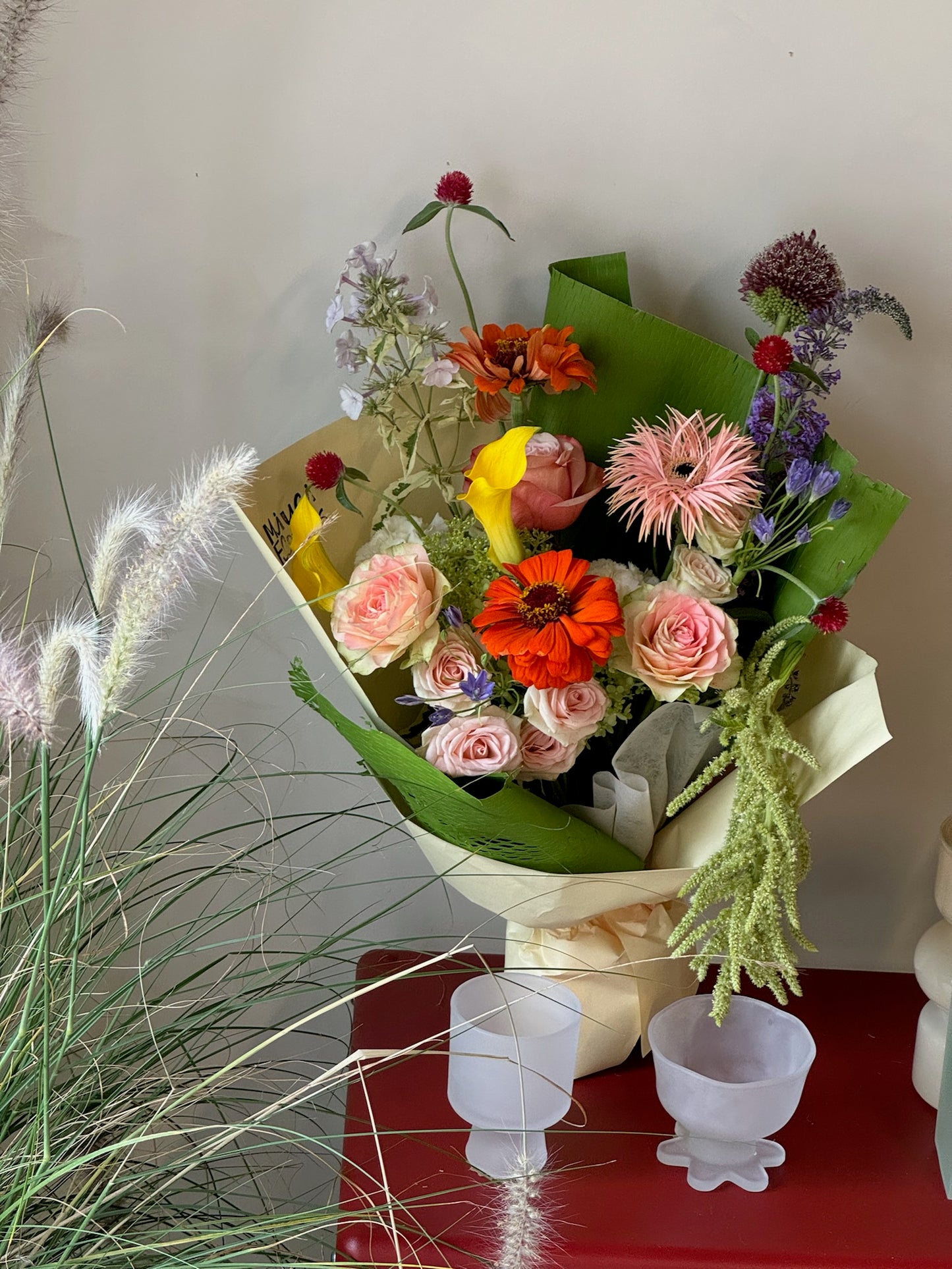 Bouquet featuring peach roses, orange zinnias, pink spider gerbera daisies, yellow calla lilies, blue delphiniums, green amaranthus, and deep red scabiosa pods. The arrangement is wrapped in green and cream paper, placed on a red table with ornamental grasses in the background.