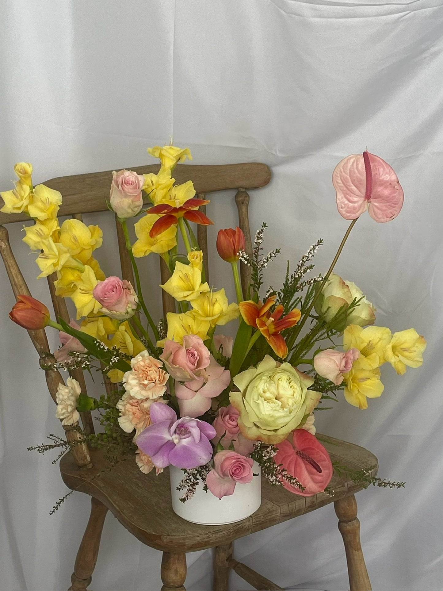 A vibrant floral arrangement displayed on a wooden chair. The arrangement features yellow gladiolus, pink roses, light orange tulips, peach carnations, pink anthurium, and purple lisianthus. The flowers are arranged in a white ceramic vase. The backdrop is a simple white fabric.