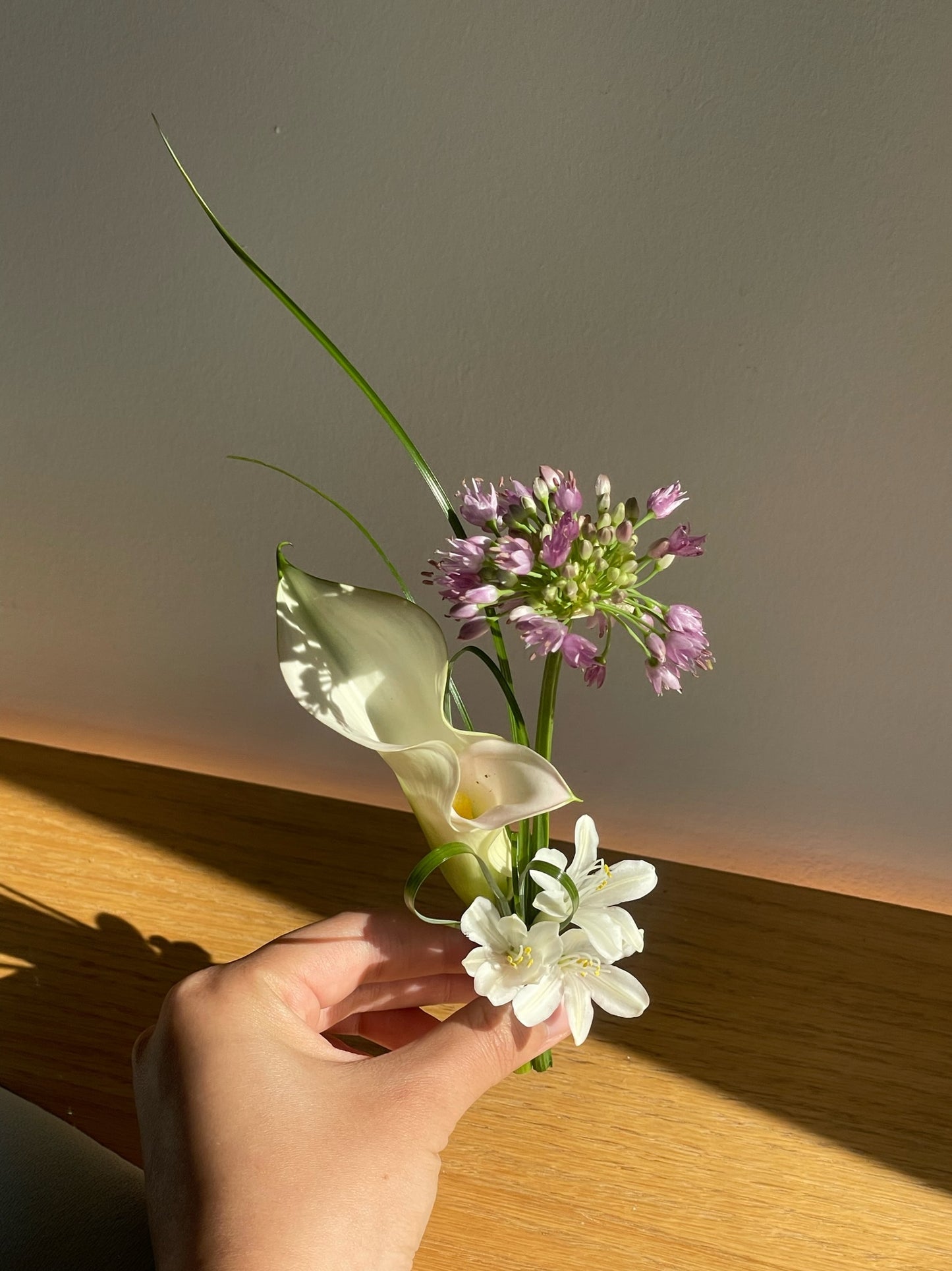A delicate boutonniere featuring a white calla lily, a small white bloom, and a cluster of purple allium flowers, accented by a single long, thin green blade of grass for a minimalist design.