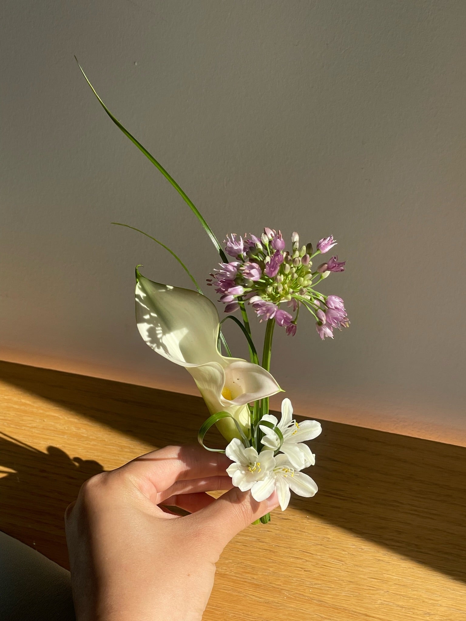 A delicate boutonniere featuring a white calla lily, a small white bloom, and a cluster of purple allium flowers, accented by a single long, thin green blade of grass for a minimalist design.