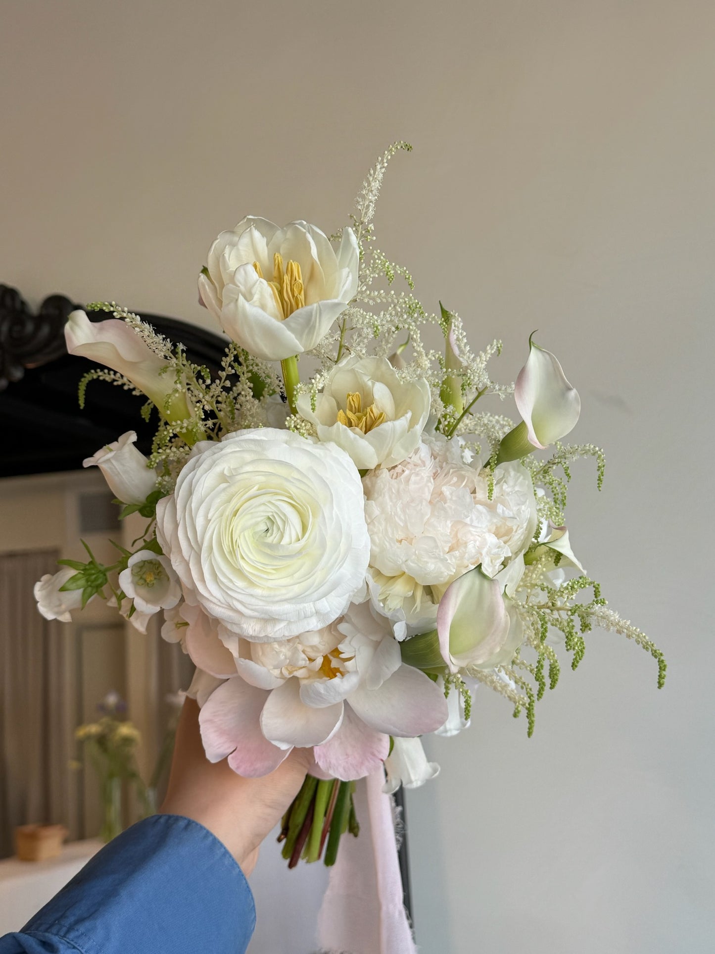 A hand-held white bridal bouquet featuring a large central ranunculus, surrounded by tulips, calla lilies, and other delicate white flowers with soft greenery accents. The bouquet is designed with a natural, elegant flow, complemented by light green foliage.