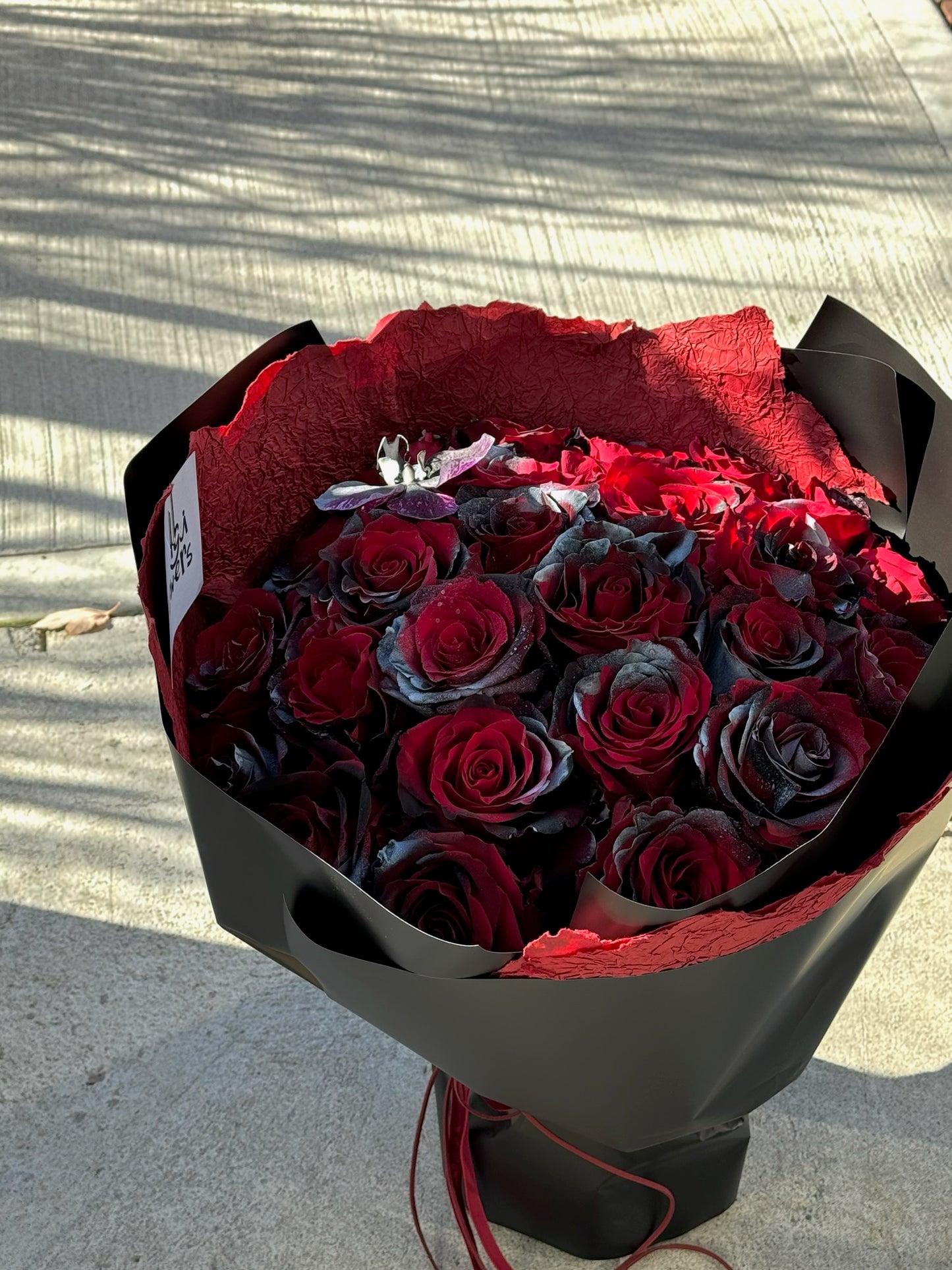 A bouquet of 24 stems of deep red roses with black-tinted petals. The bouquet is wrapped in black paper with a textured red inner lining, tied with a red ribbon. A branded card is tucked into the bouquet, and it is displayed outdoors on a concrete surface under natural sunlight.