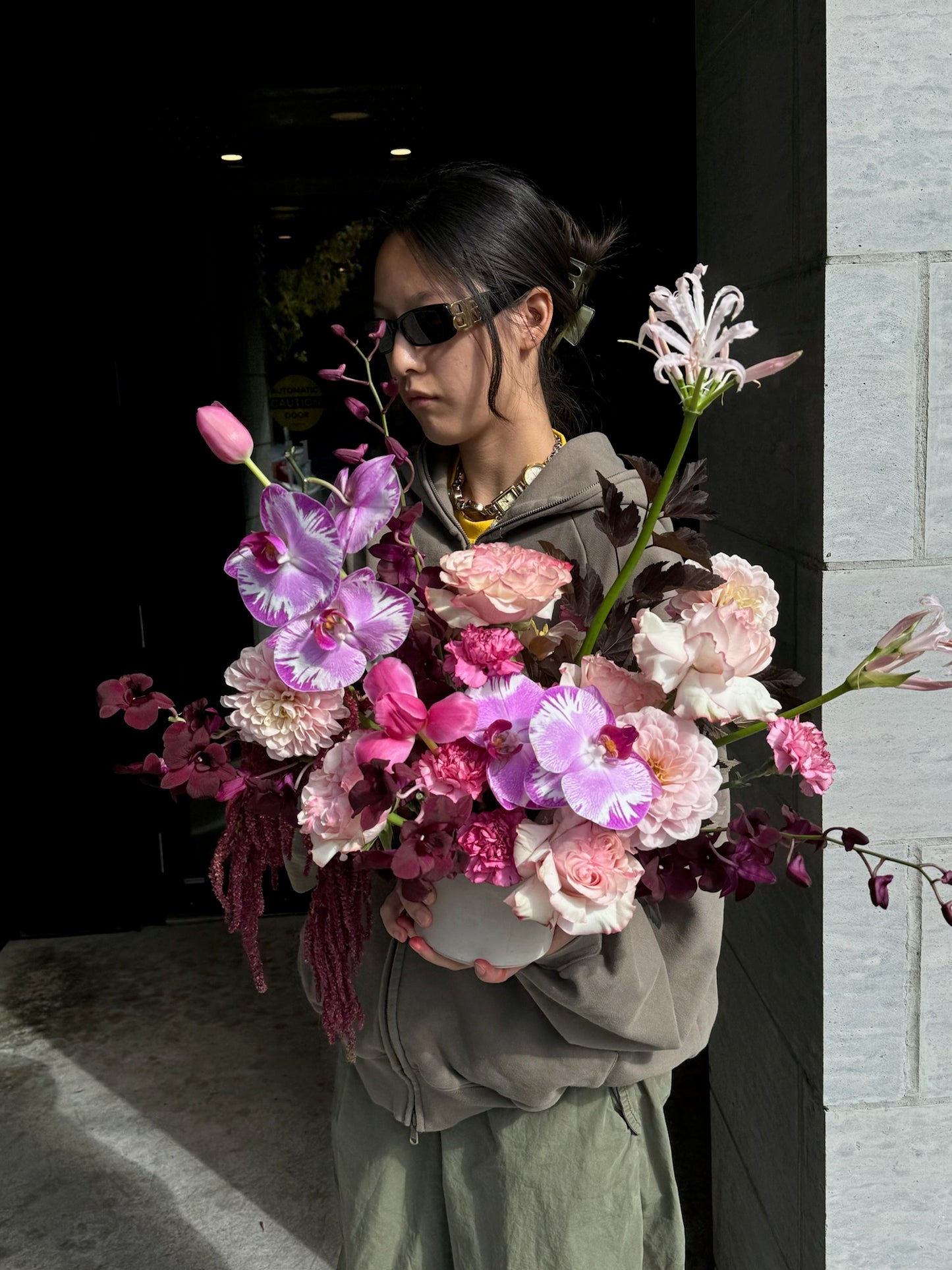 Person holding a vibrant bouquet featuring purple lisianthus, pink roses, carnations, and greenery in a white vase, standing outside in natural light.