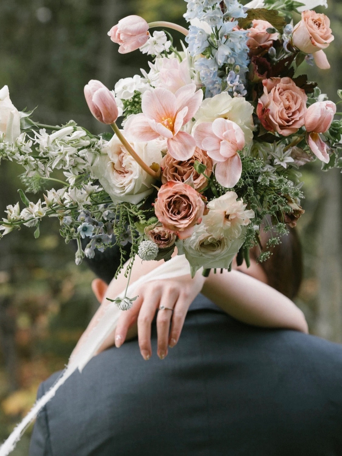 A garden-style bridal bouquet featuring blush, peach, cream, and blue flowers with greenery, held by a bride embracing a groom outdoors in a natural setting.