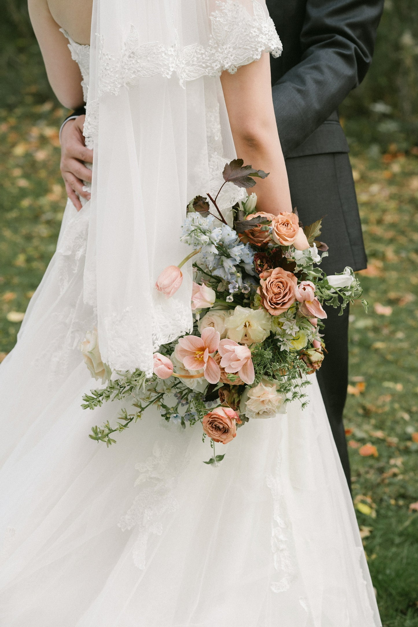 A garden-style bridal bouquet with peach, blush, and cream flowers, accented by greenery and blue blooms, held by a bride in a white lace dress with a groom in a dark suit, photographed outdoors on a grassy background.