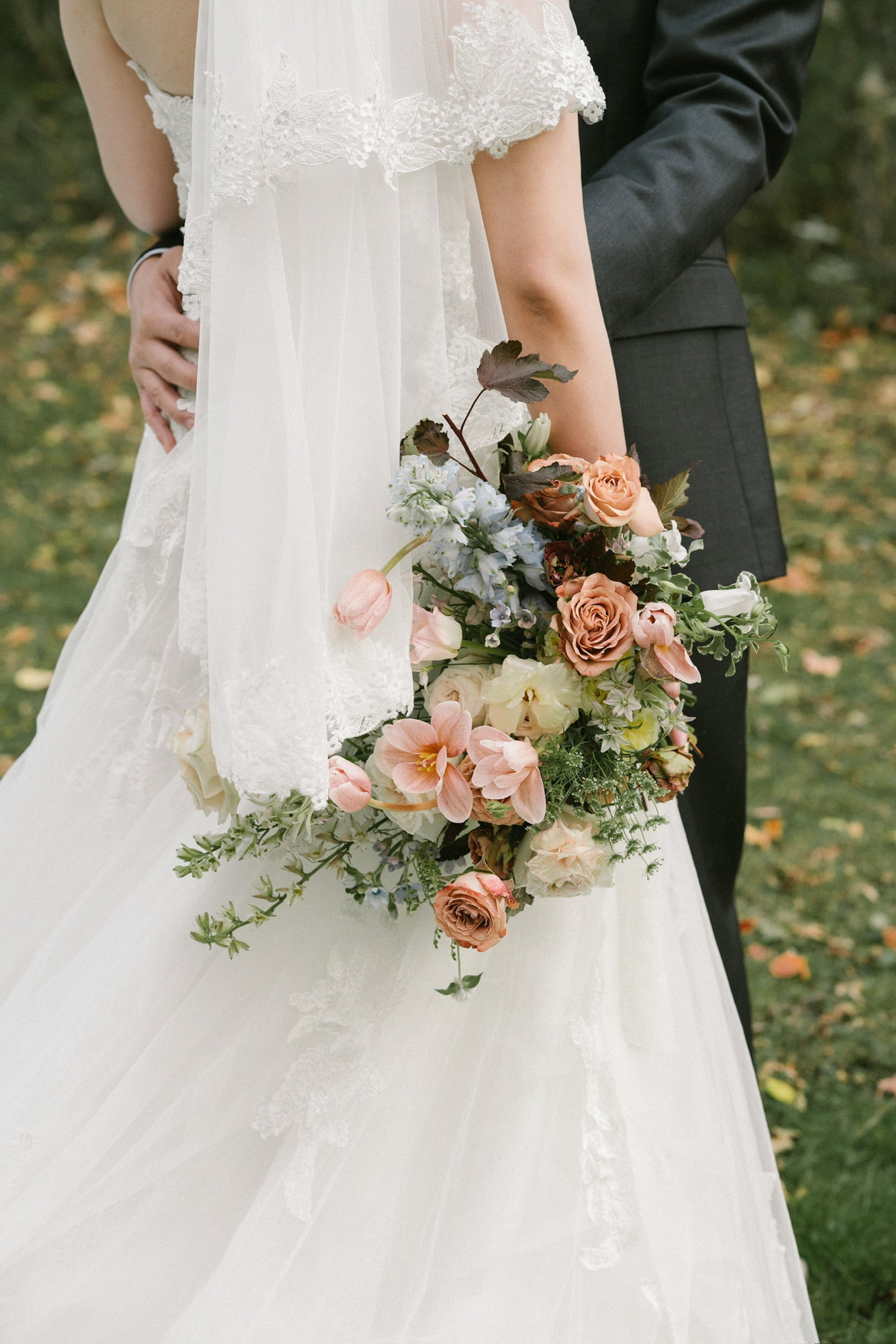 A garden-style bridal bouquet with peach, blush, and cream flowers, accented by greenery and blue blooms, held by a bride in a white lace dress with a groom in a dark suit, photographed outdoors on a grassy background.