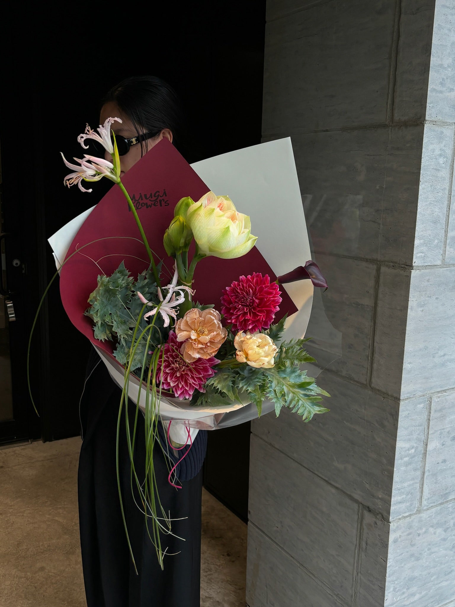 A bouquet featuring yellow tulips, maroon dahlias, peach carnations, and greenery, accented with cascading floral elements. The bouquet is wrapped in white and maroon paper, held by a person standing in front of a stone wall, partially in shadow.