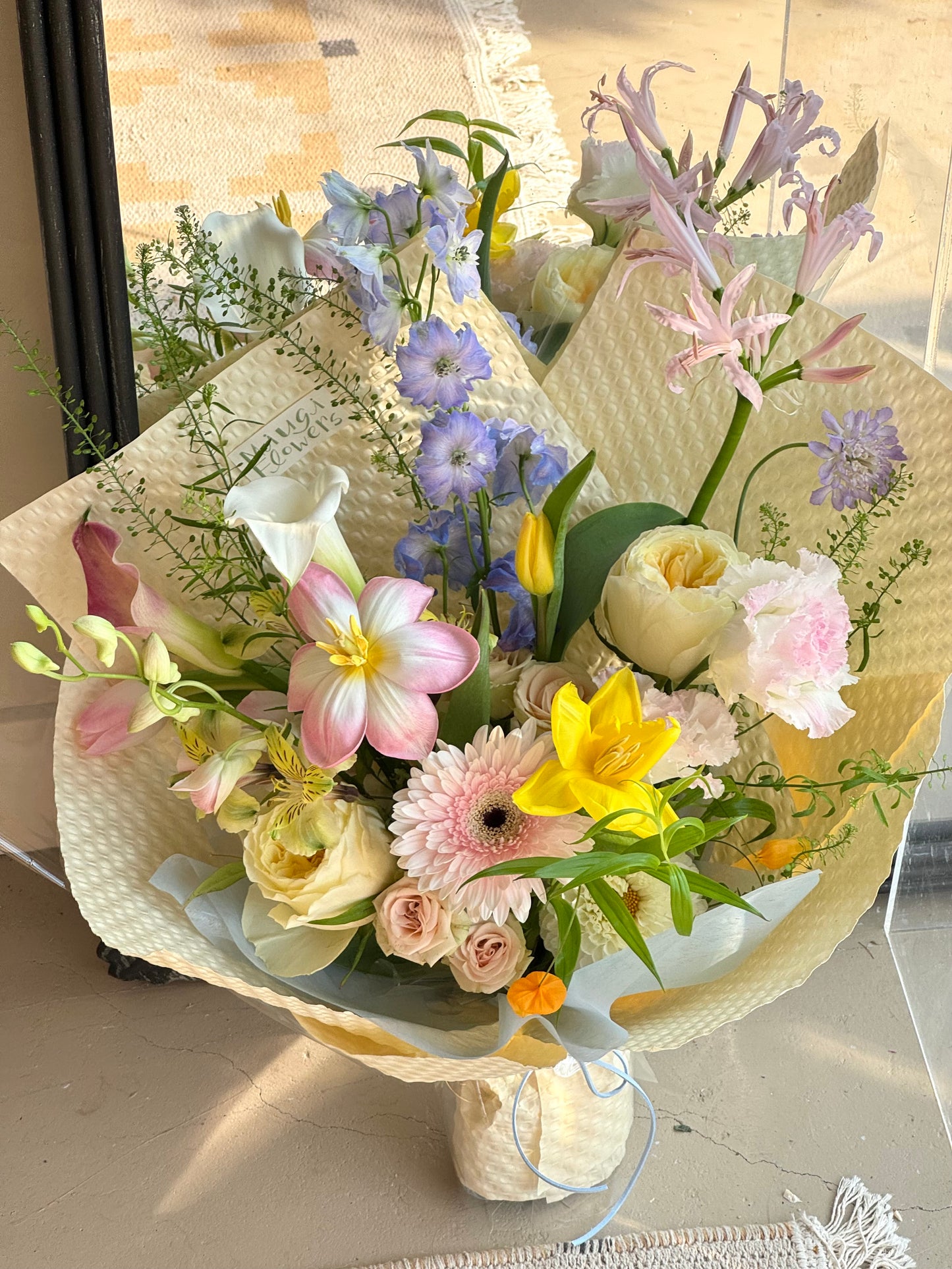 Colorful floral bouquet featuring pink, yellow, and purple flowers, including lilies, gerbera daisies, roses, and delphiniums, arranged in a white woven wrap with greenery accents.