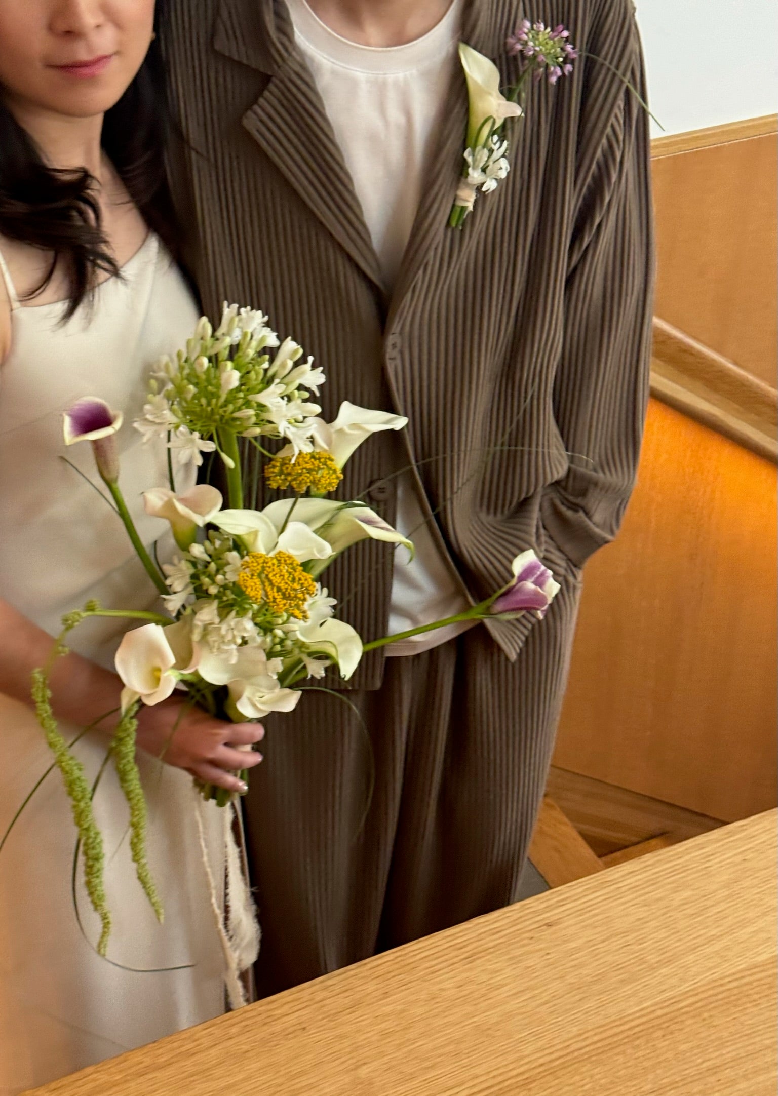A bride and groom standing together, with the bride holding a bouquet of white calla lilies, yellow yarrow, purple and white blooms, and delicate greenery. The groom is wearing a boutonnière with matching flowers.