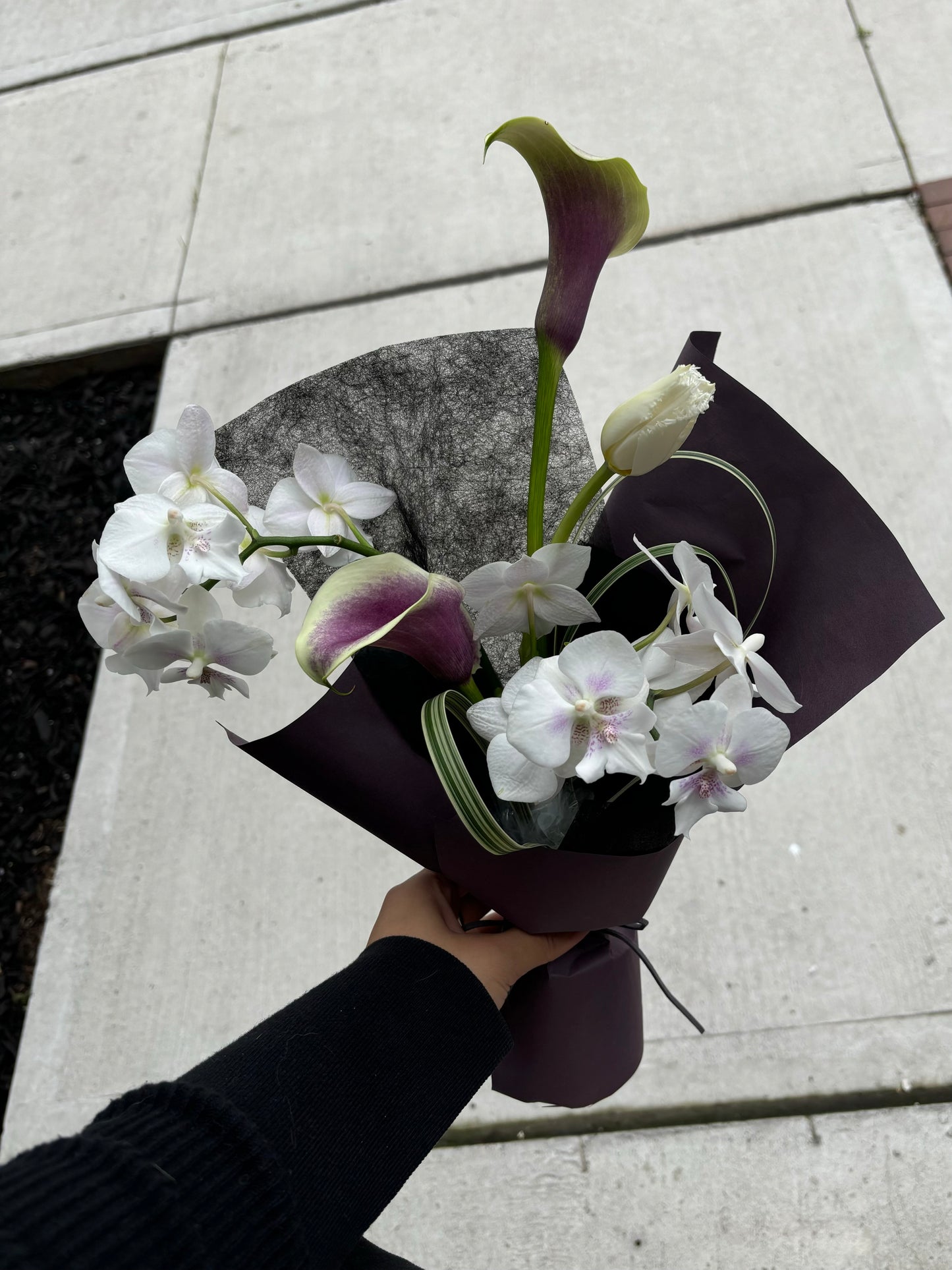A hand holding a small bouquet featuring purple calla lilies and white mini orchids, wrapped in dark brown paper, photographed outdoors against a concrete sidewalk.