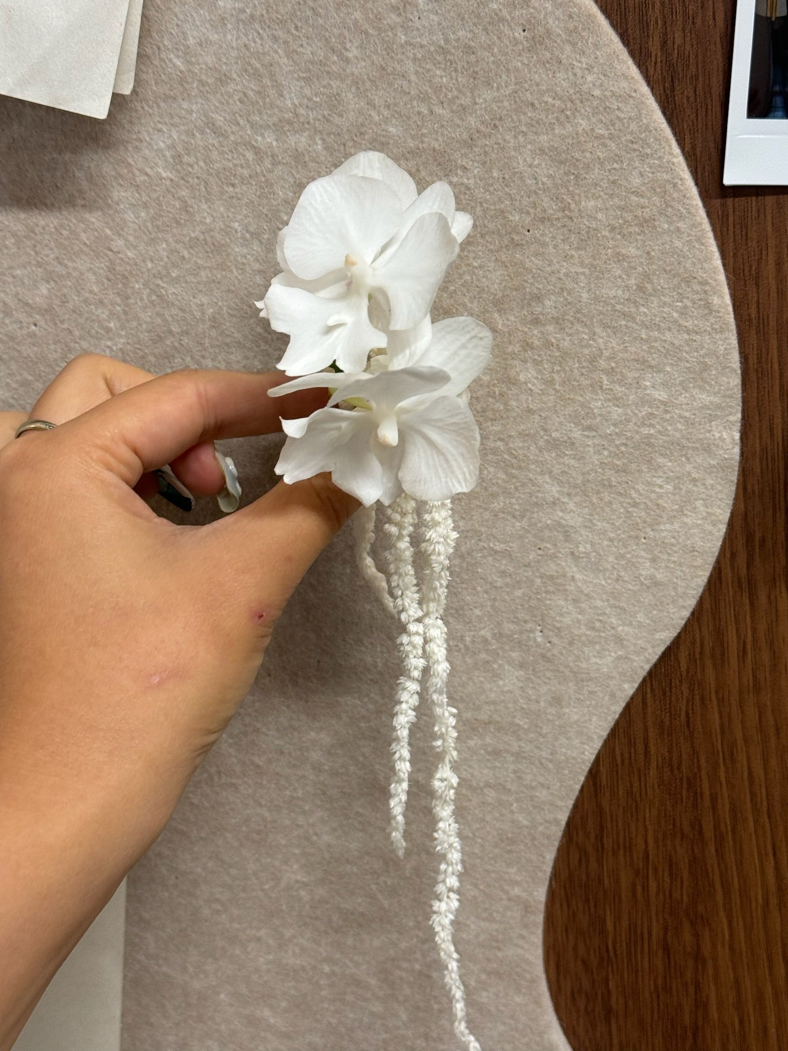 Hand holding a boutonniere featuring a white mini orchid and white dried amaranthus set on a beige felt surface.