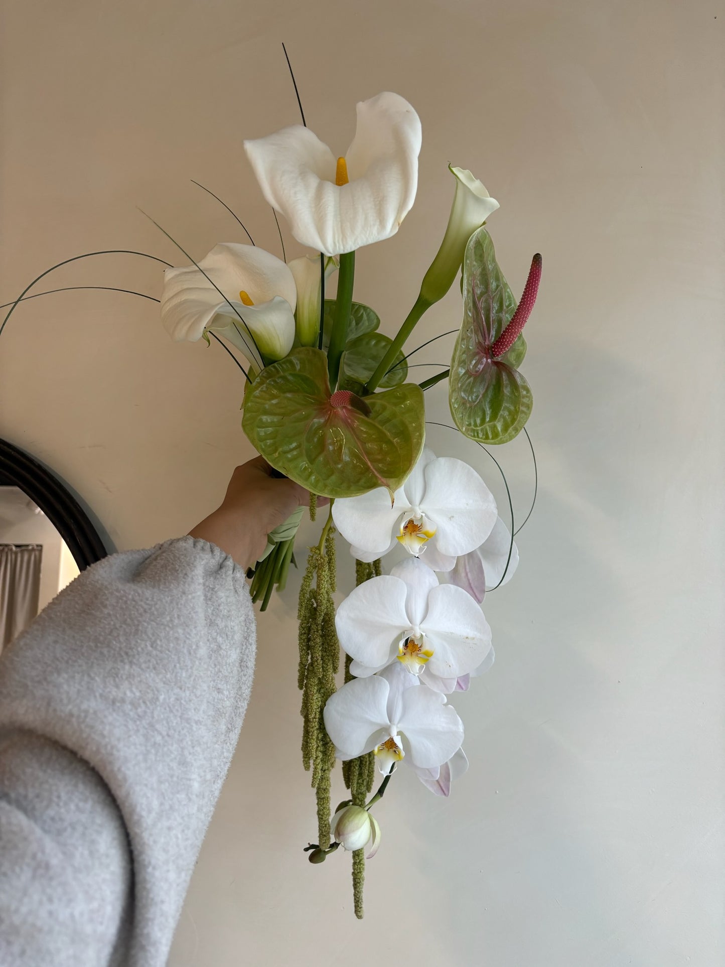 A bridal bouquet featuring white calla lilies, white orchids, and green anthurium leaves, accented by delicate greenery. The bouquet is held by a person wearing a light gray sweater, displayed against a neutral indoor background with soft natural lighting.