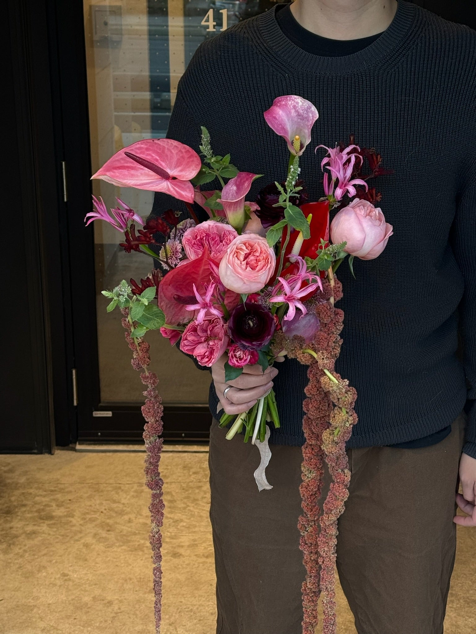 A bridal bouquet featuring pink garden roses, pink and red anthurium, ranunculus, gloriosa lilies, and dark accents, paired with cascading amaranthus and lush greenery.