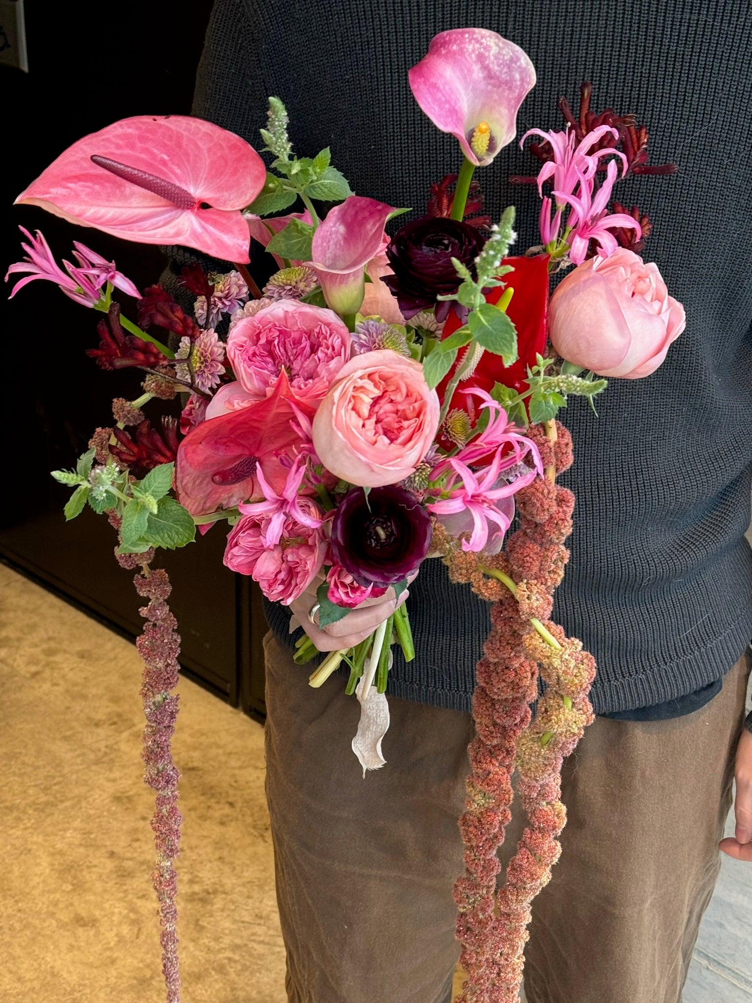 A close-up of a pink bridal bouquet featuring lush garden roses, anthurium, ranunculus, gloriosa lilies, and trailing red amaranthus.