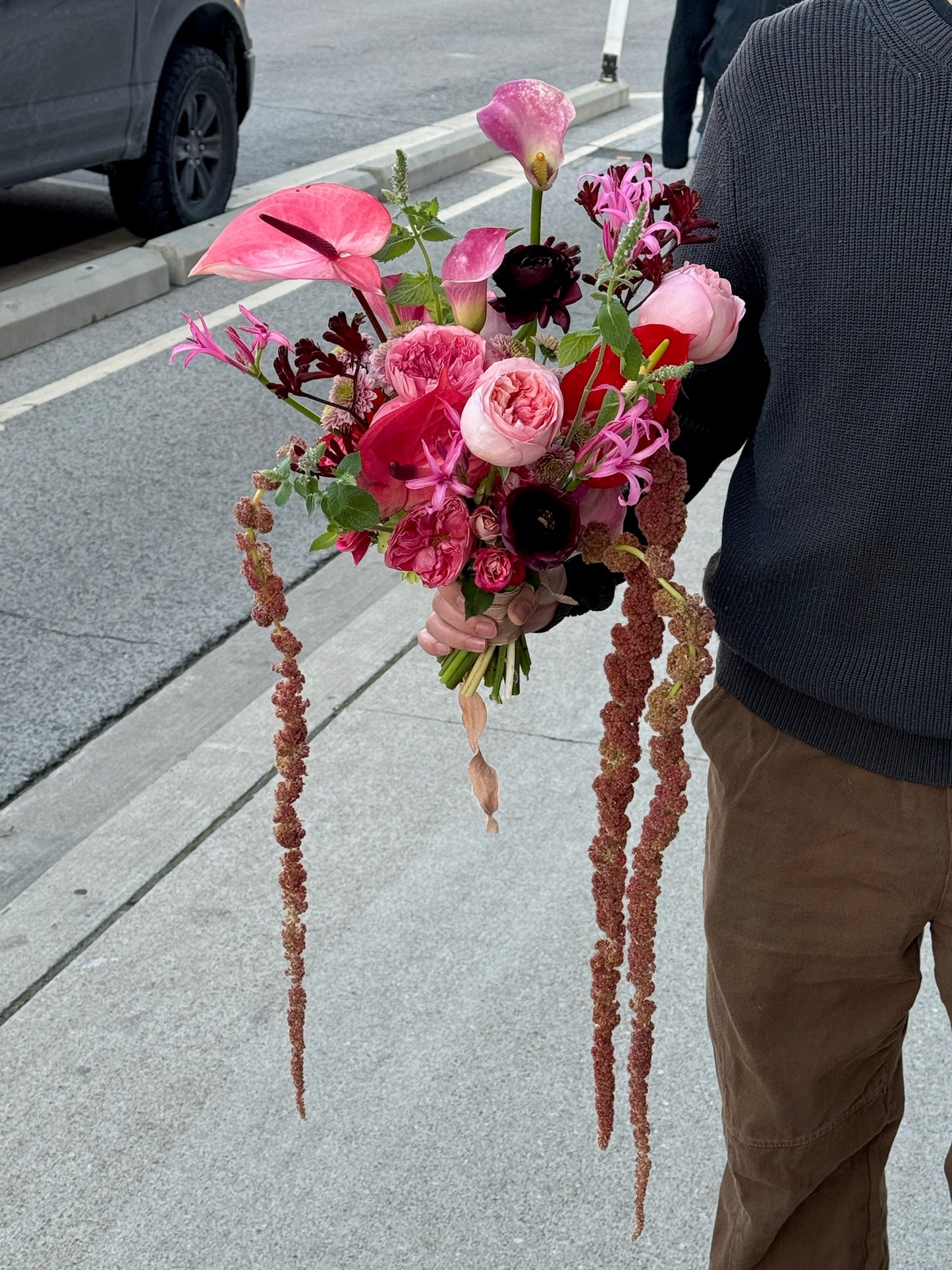 A bridal bouquet featuring pink roses, anthurium, pink garden roses, purple ranunculus, and gloriosa lilies, accented with trailing amaranthus and lush greenery, creating a romantic and cascading design.