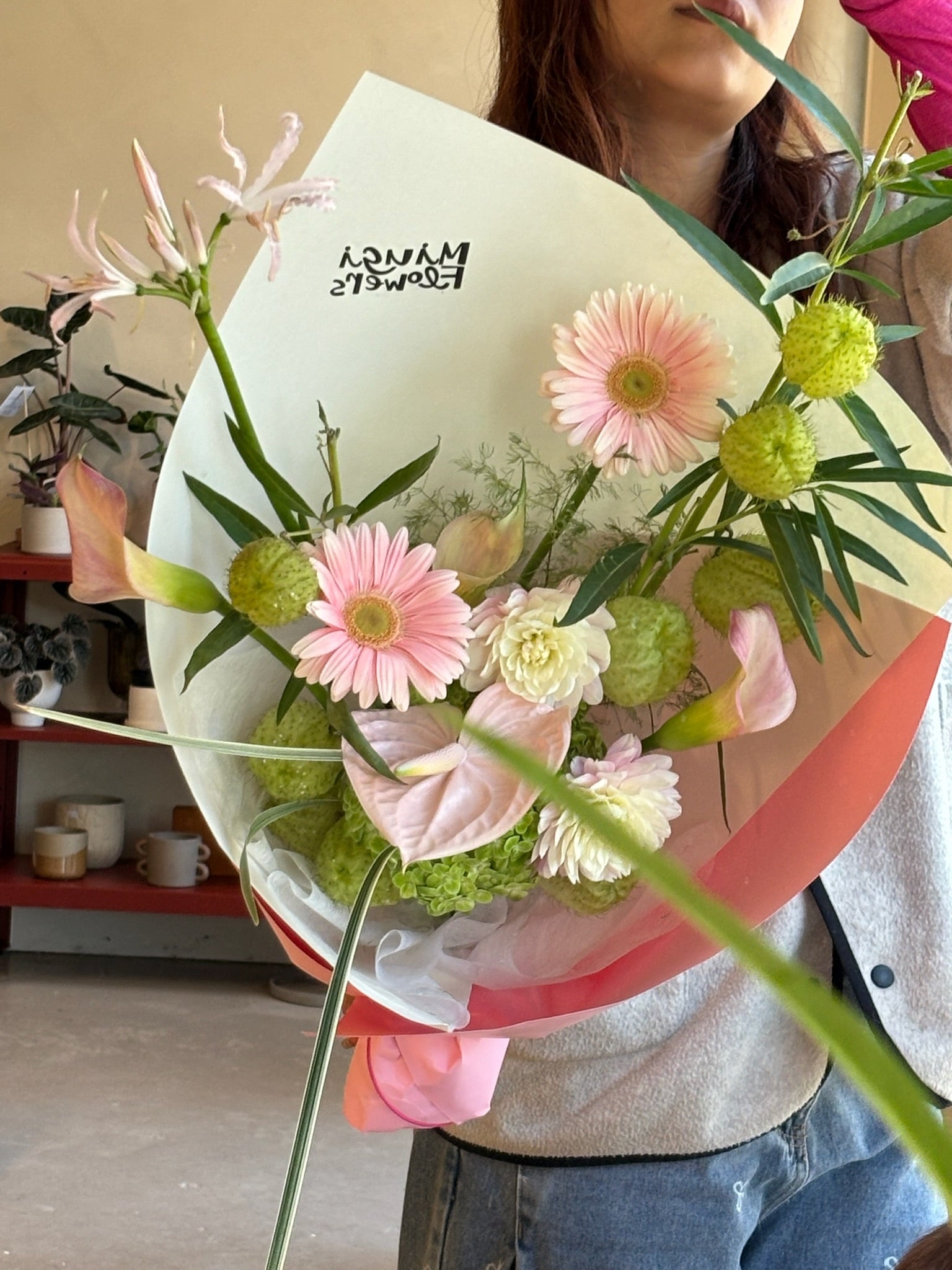 A bouquet featuring soft pink gerbera daisies, green dianthus, white lisianthus, and delicate pink nerines, arranged with green foliage. The flowers are wrapped in white and soft pink paper, held by a person with “Miugi Flowers” branding visible on the wrapping.
