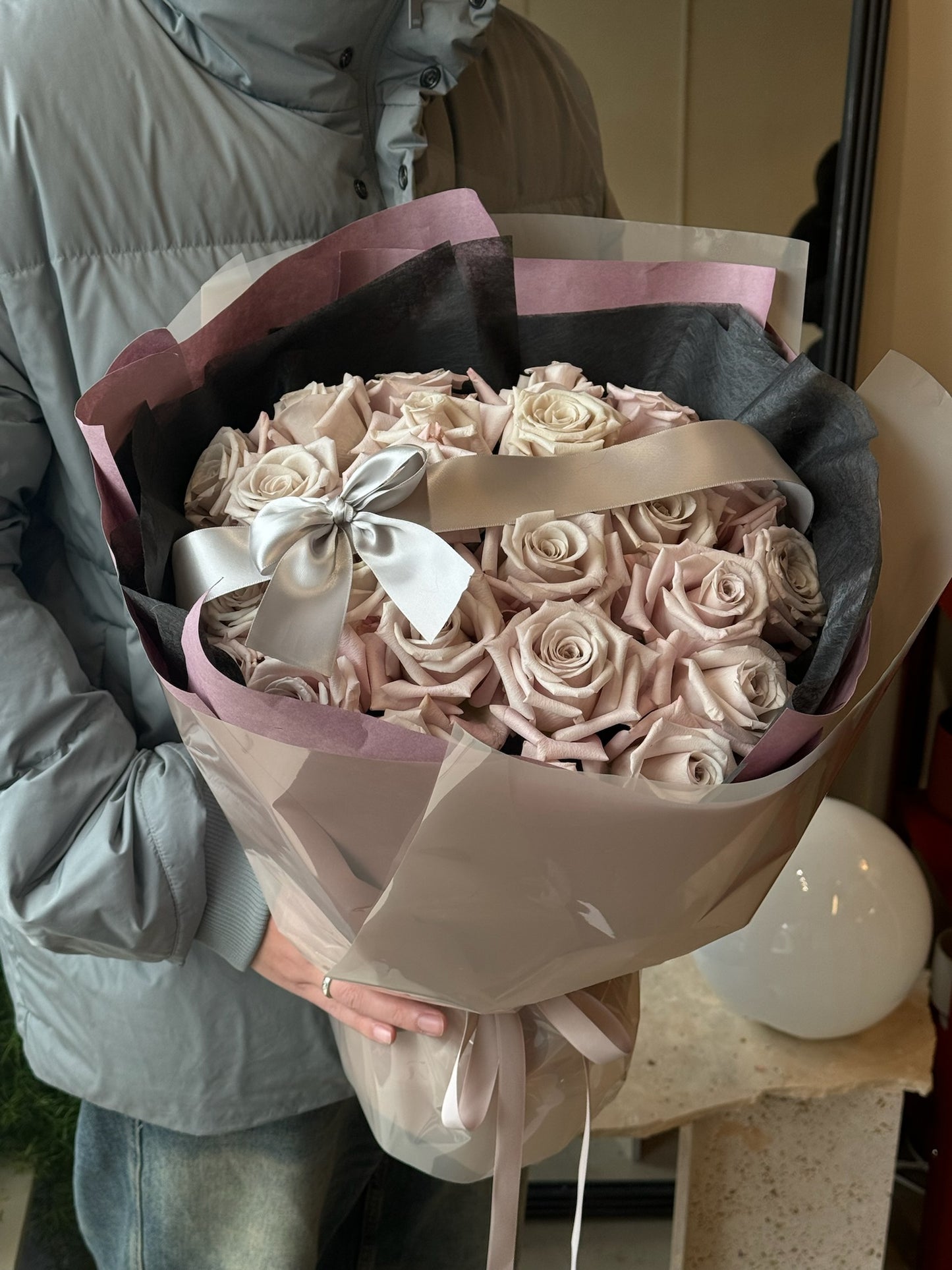 A bouquet of soft beige manta roses arranged in an elegant style, wrapped in layered pink and gray paper and tied with a silver ribbon. The bouquet is being held by a person wearing a light gray jacket, displayed indoors with a neutral background and decorative elements.