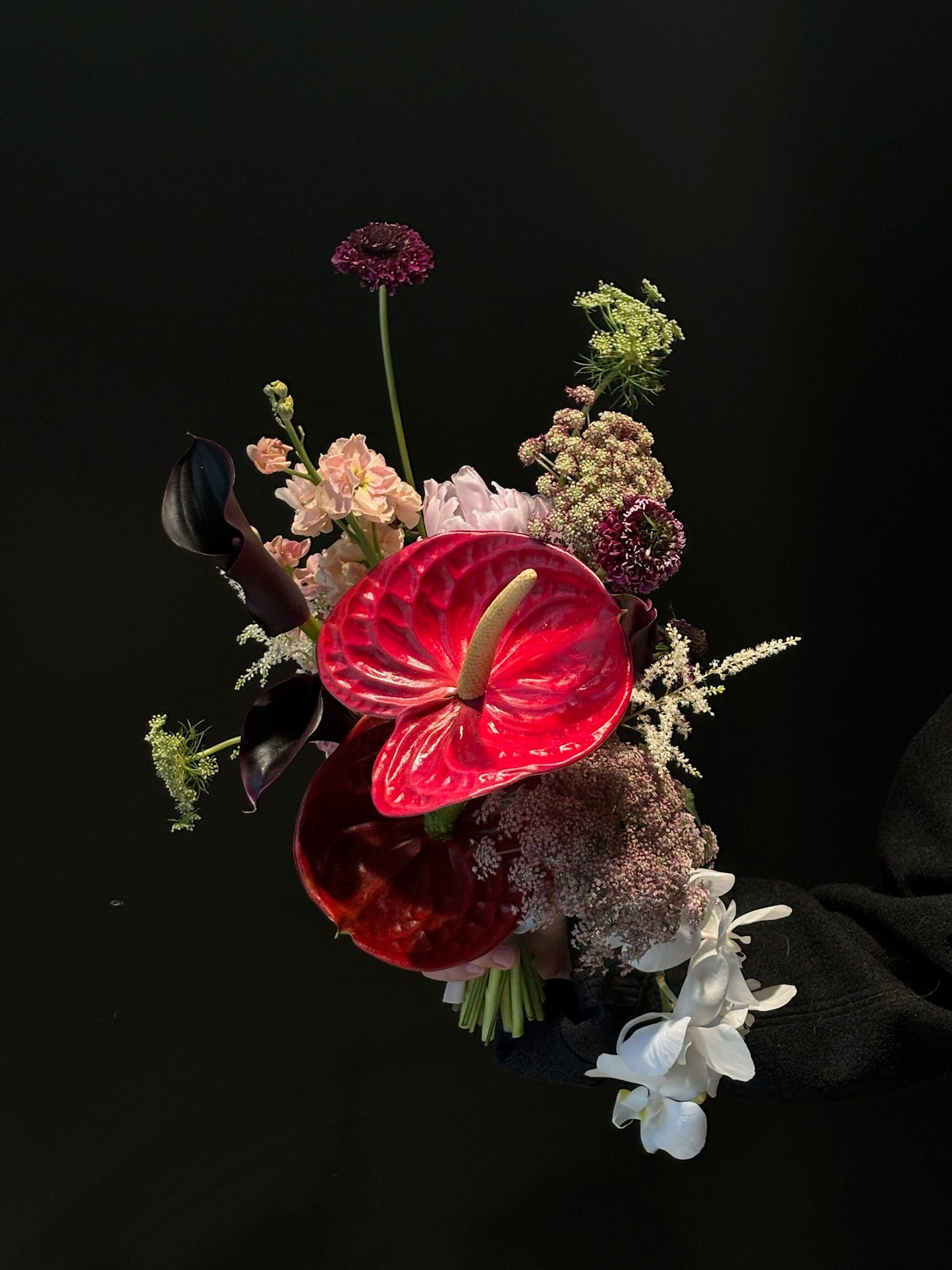 A bridal bouquet featuring a vibrant red anthurium, black calla lilies, white orchids, and a mix of textured flowers and greenery. The arrangement is displayed against a dark black background, highlighting the bold and elegant colors and textures of the blooms.