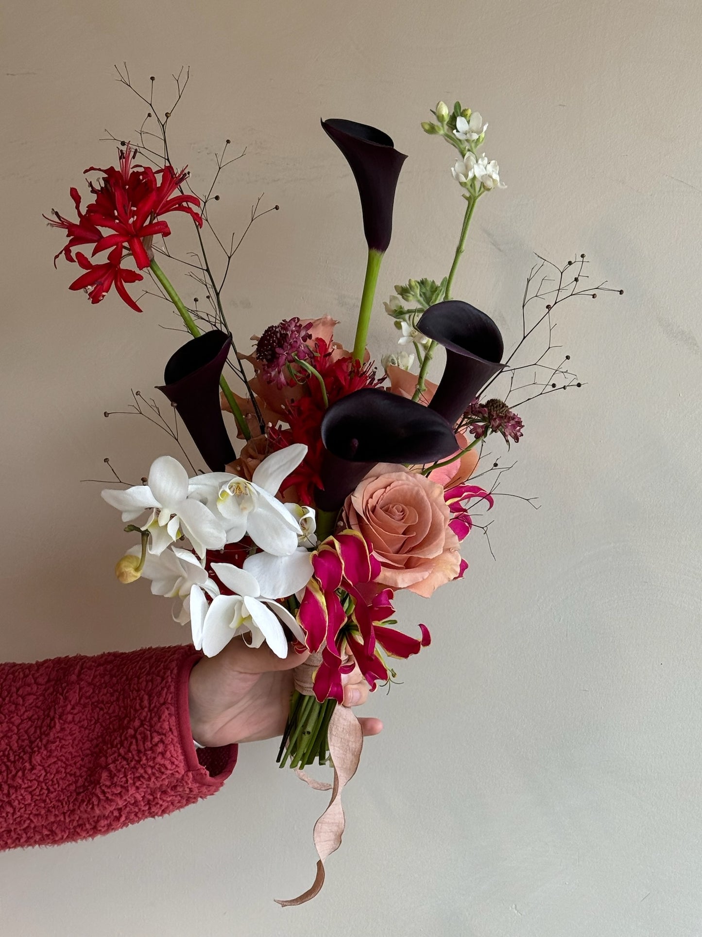 A modern bouquet featuring deep black calla lilies, red gloriosa lilies, soft peach roses, white orchids, and delicate filler flowers, arranged with a touch of whimsy and elegance.
