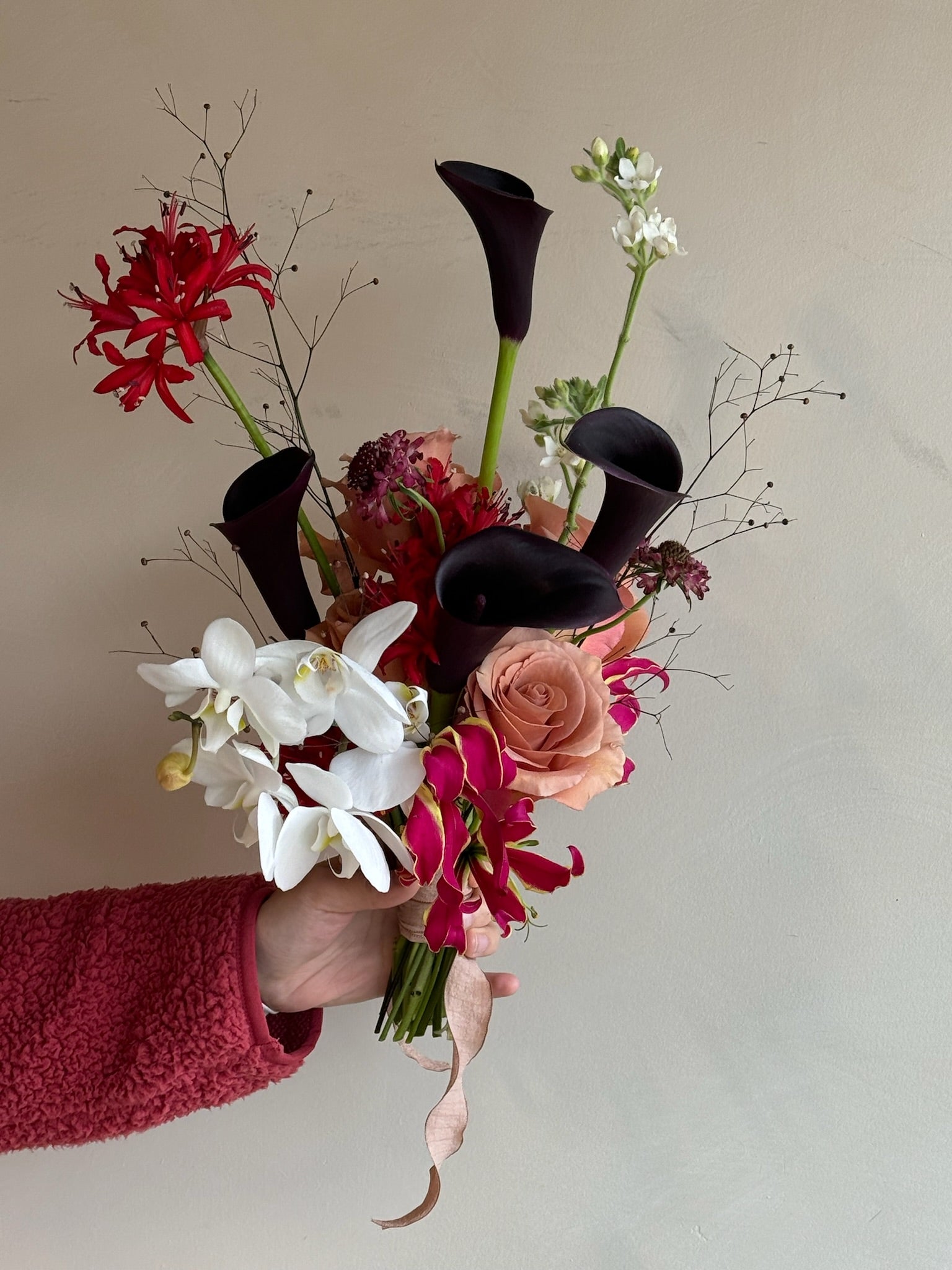 A modern bouquet featuring deep black calla lilies, red gloriosa lilies, soft peach roses, white orchids, and delicate filler flowers, arranged with a touch of whimsy and elegance.