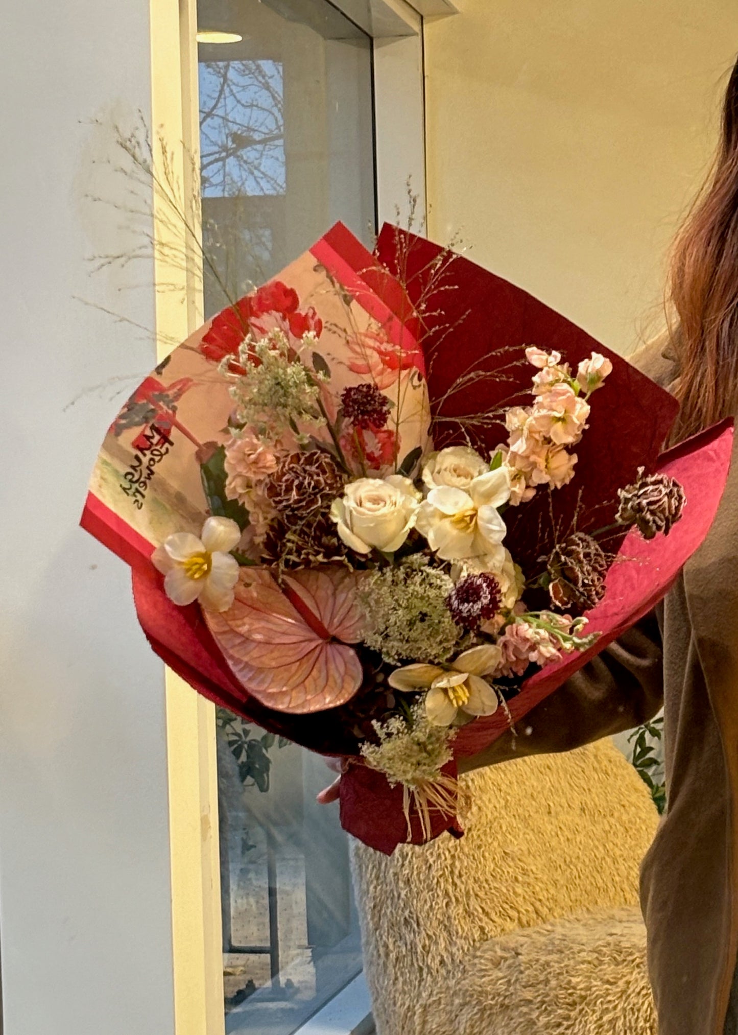A close-up of a warm-toned bouquet featuring white roses, pink anthurium, dried flowers, and delicate blooms in earthy shades. The bouquet is wrapped in red and beige paper, held by a person in an indoor setting with natural light and a soft background.