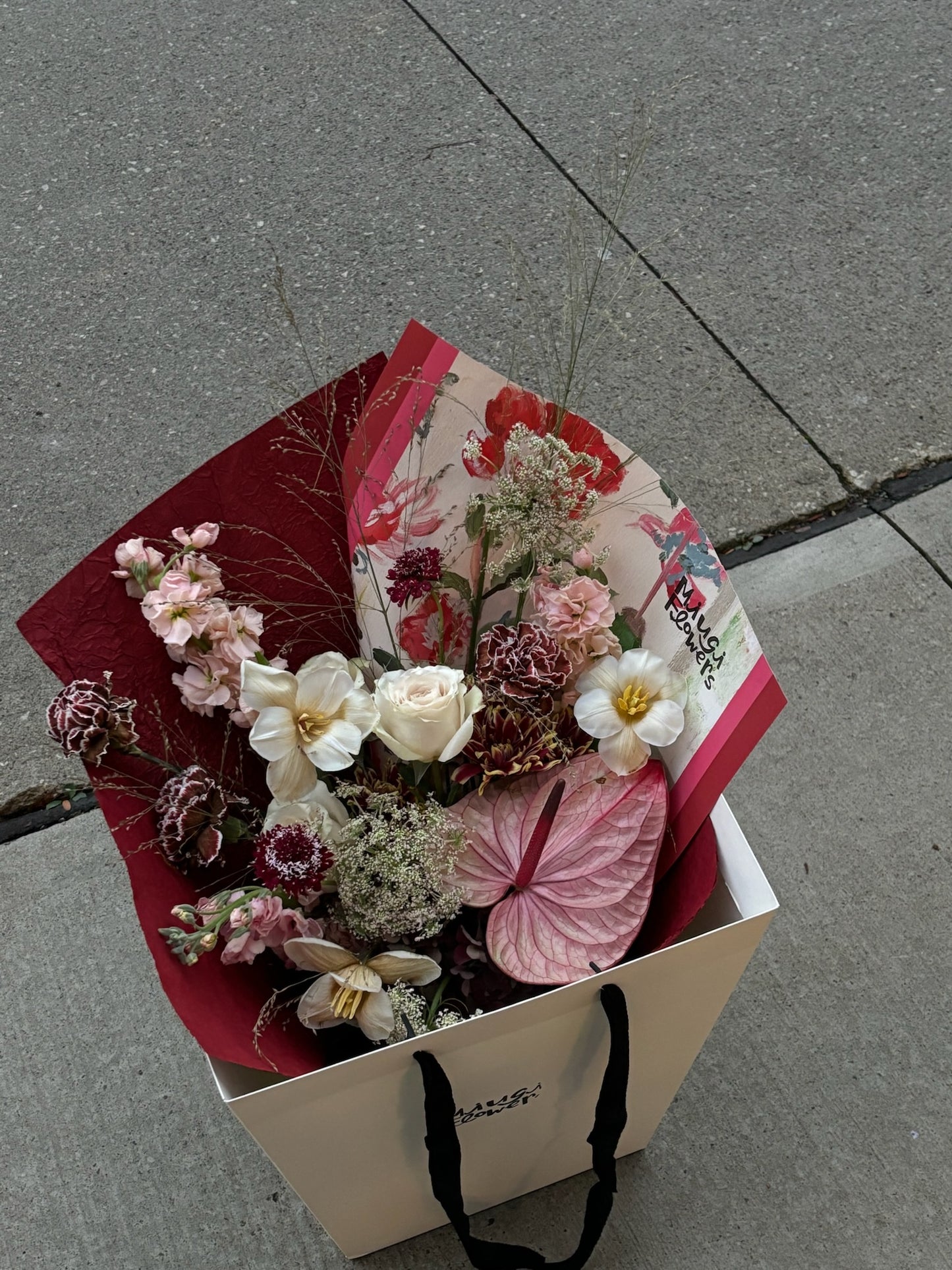 A bouquet featuring white roses, pink anthurium, dried flowers, and other delicate blooms in soft and muted tones. The arrangement is wrapped in red and beige paper, placed in a cream-colored gift bag with black handles. It is displayed outdoors on a concrete surface.