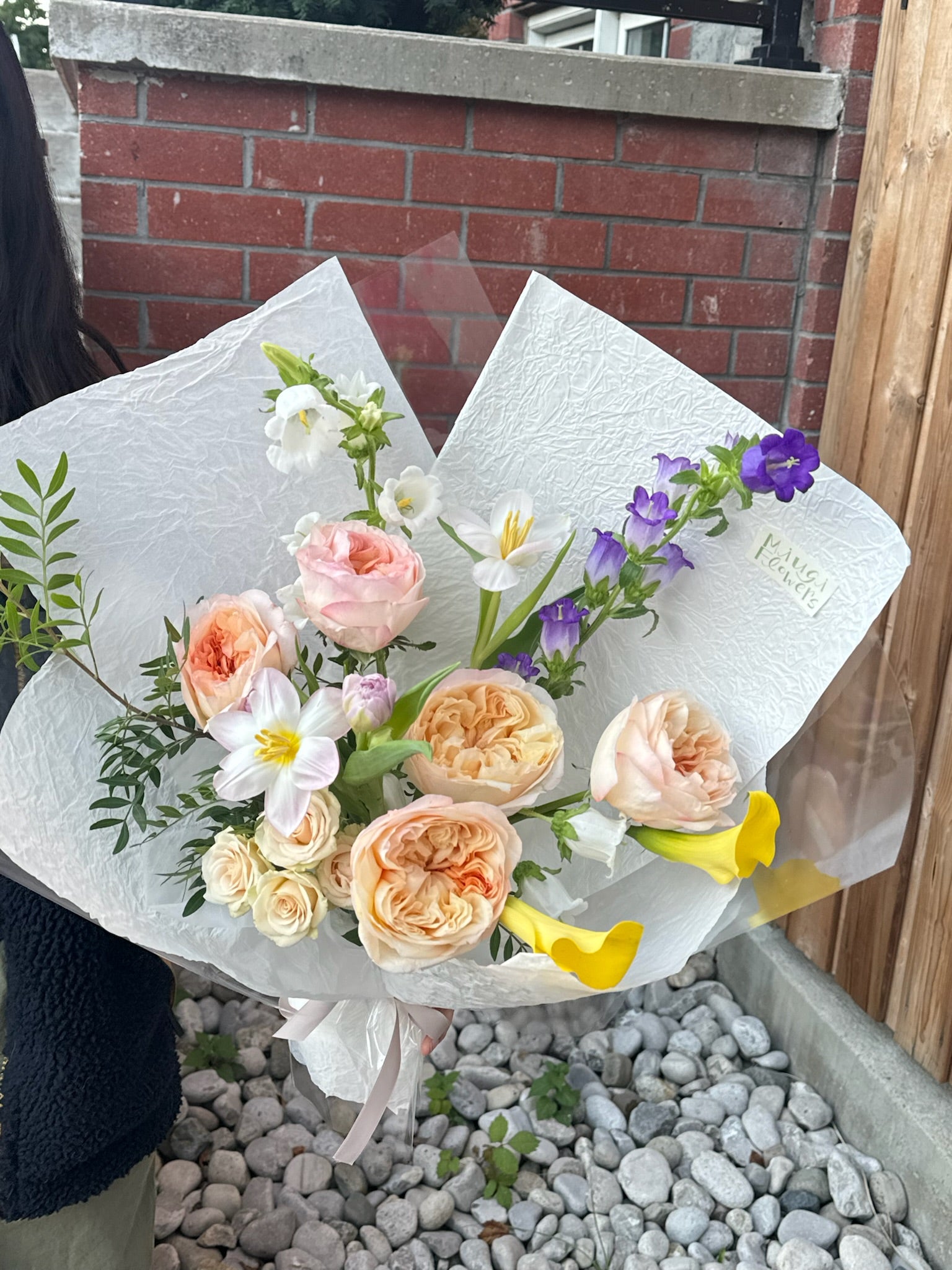 A bouquet featuring soft blush peach garden roses, white lisianthus, purple delphinium, and yellow calla lilies, wrapped in textured white paper. The arrangement is displayed outdoors near a wooden fence and a brick wall.