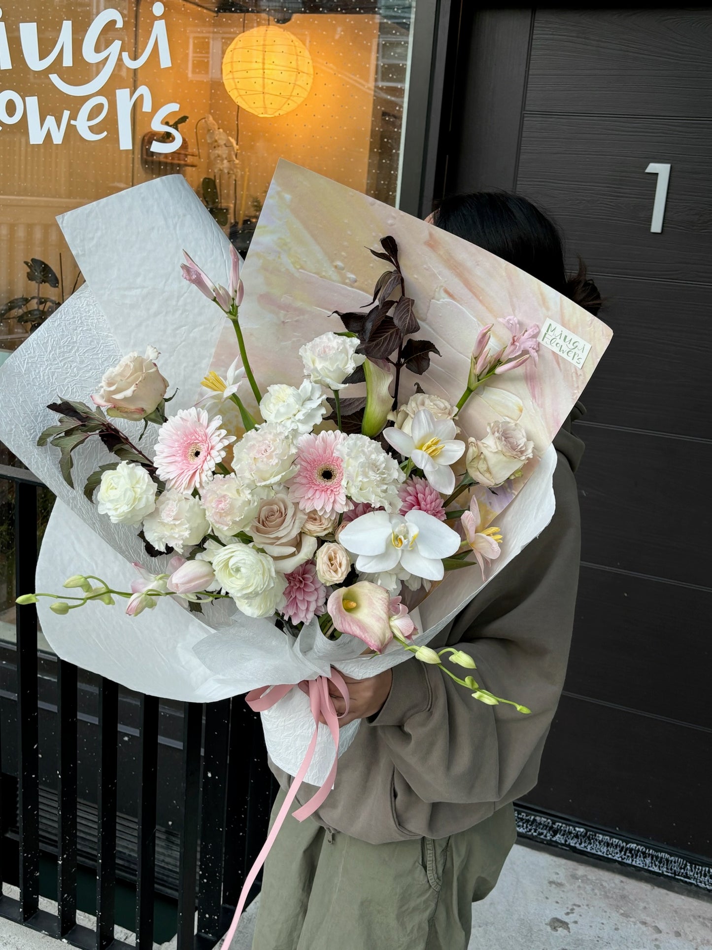 Person holding a large pastel-toned bouquet featuring roses, gerbera daisies, carnations, and lisianthus wrapped in white paper with dark foliage, standing outside Miugi Flowers shop.