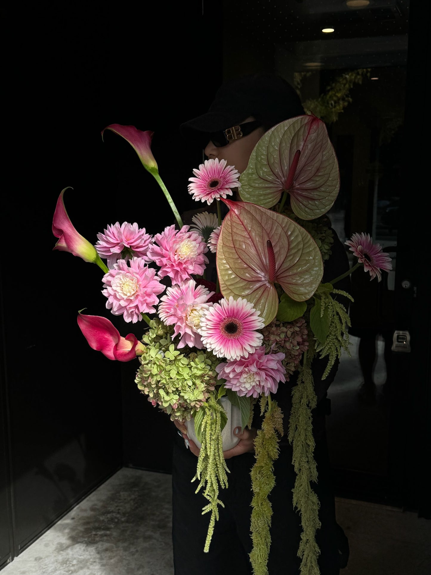 Vase Arrangement featuring pistachio anthuriums, gerbera daisies, calla lilies, and hanging amaranthus, held by a person in a dark setting.