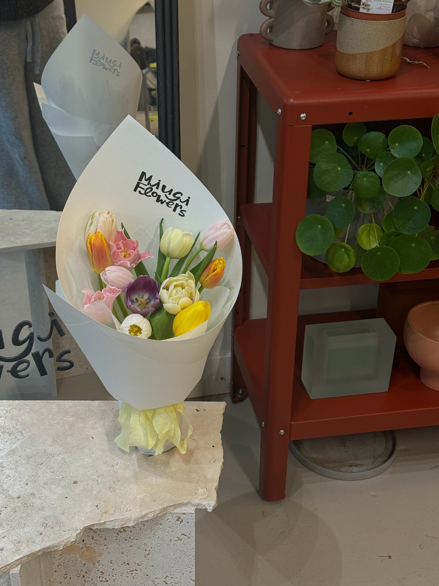 A colorful tulip bouquet wrapped in white paper, labeled with “Miugi Flowers.” The arrangement features a mix of tulips in yellow, pink, purple, and orange shades. The bouquet sits on a textured stone surface with a red shelf and green potted plant in the background.