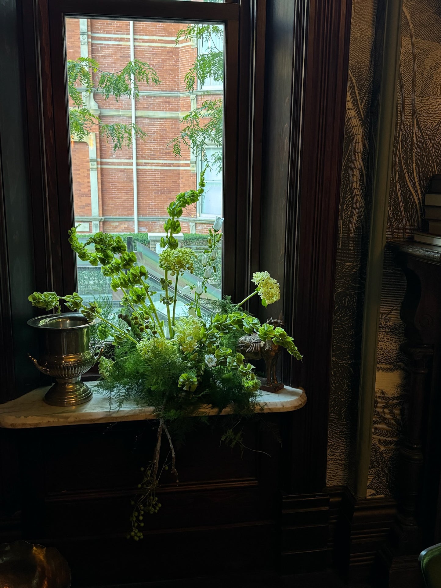 A lush green floral arrangement placed on a wooden windowsill, featuring ferns, trailing vines, and green hydrangeas. The arrangement is set beside a silver urn-style vase, with a view of a brick building and outdoor greenery through the window. The setting is in a dimly lit room with dark wood paneling, creating a cozy and vintage atmosphere.