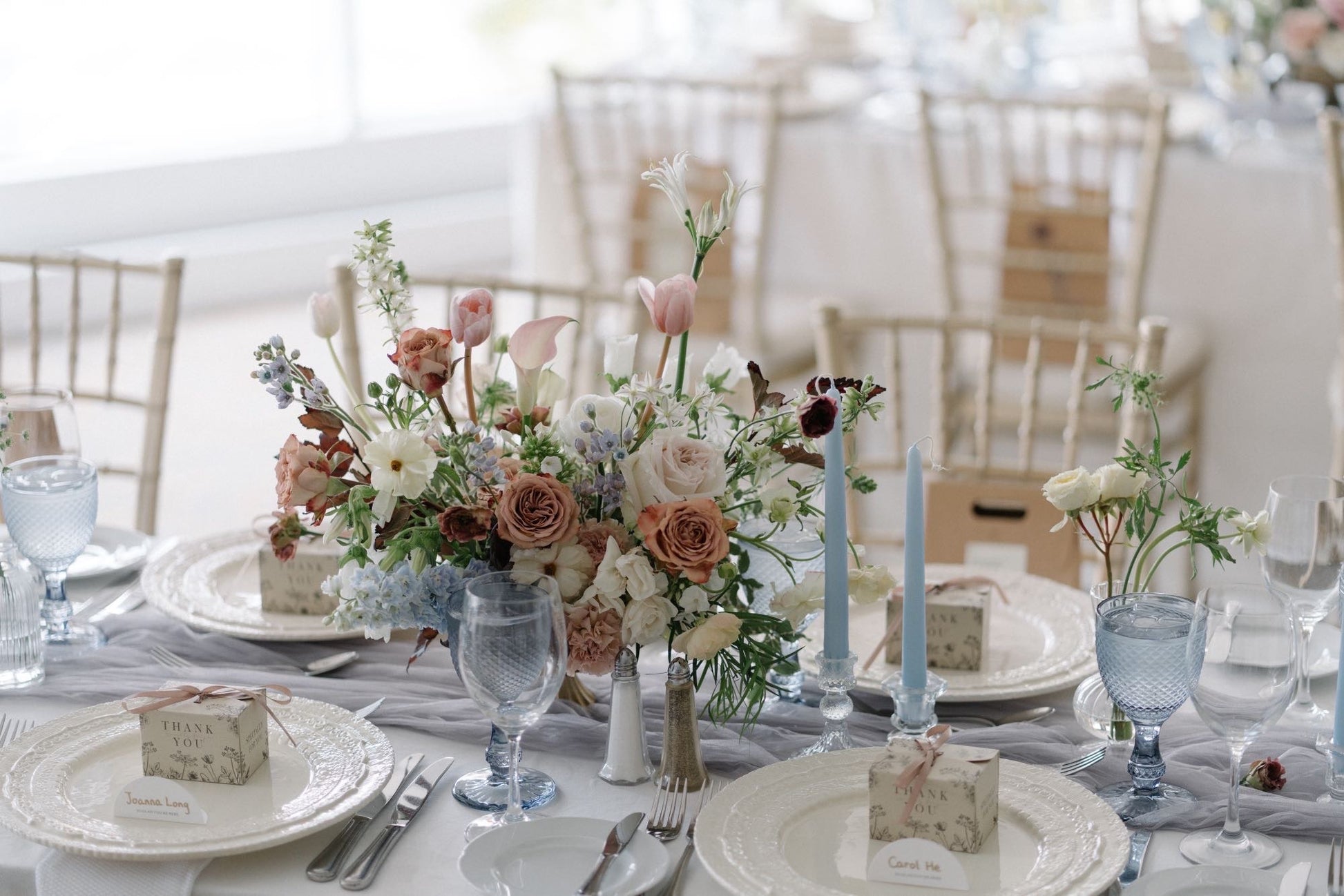 A formal dining table featuring a floral centerpiece with blush roses, white ranunculus, light pink tulips, blue hydrangeas, and small white flowers. The table is set with white plates, silver cutlery, light blue water goblets, and two blue taper candles in glass holders. Each plate has a small cream-colored gift box and a name card. White chairs and a neutral background are visible.