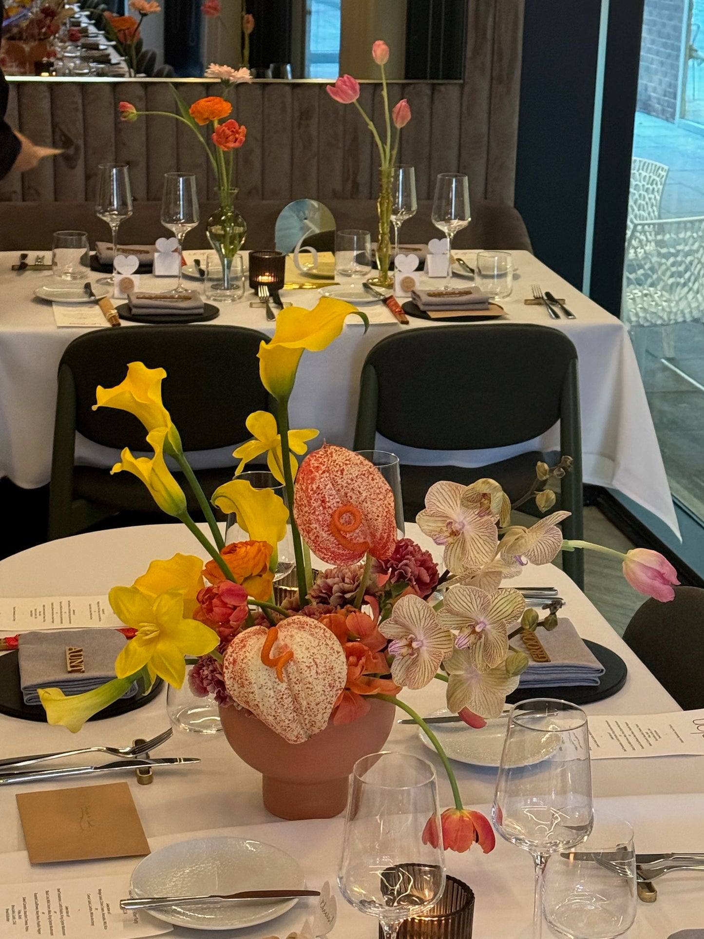 A floral arrangement in a terracotta pot featuring yellow calla lilies, orange pincushion protea, pink tulips, and light pink orchids. The arrangement is placed on a formal dining table set with wine glasses, white napkins, and silver cutlery. The background includes additional tables with similar setups.