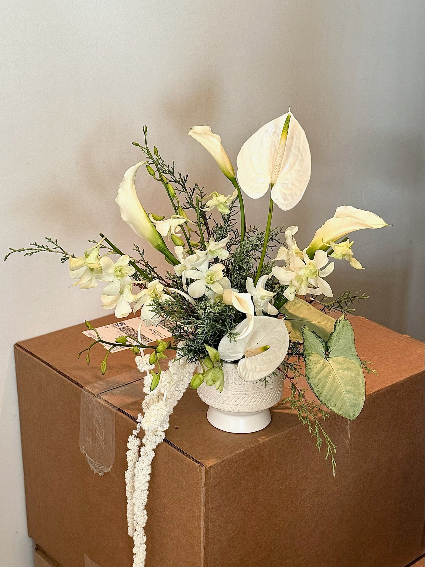 A floral arrangement in a white vase featuring white anthuriums, white calla lilies, and white dendrobium orchids. The arrangement is accented with soft greenery and trailing white amaranthus, placed on top of a cardboard box against a neutral background.