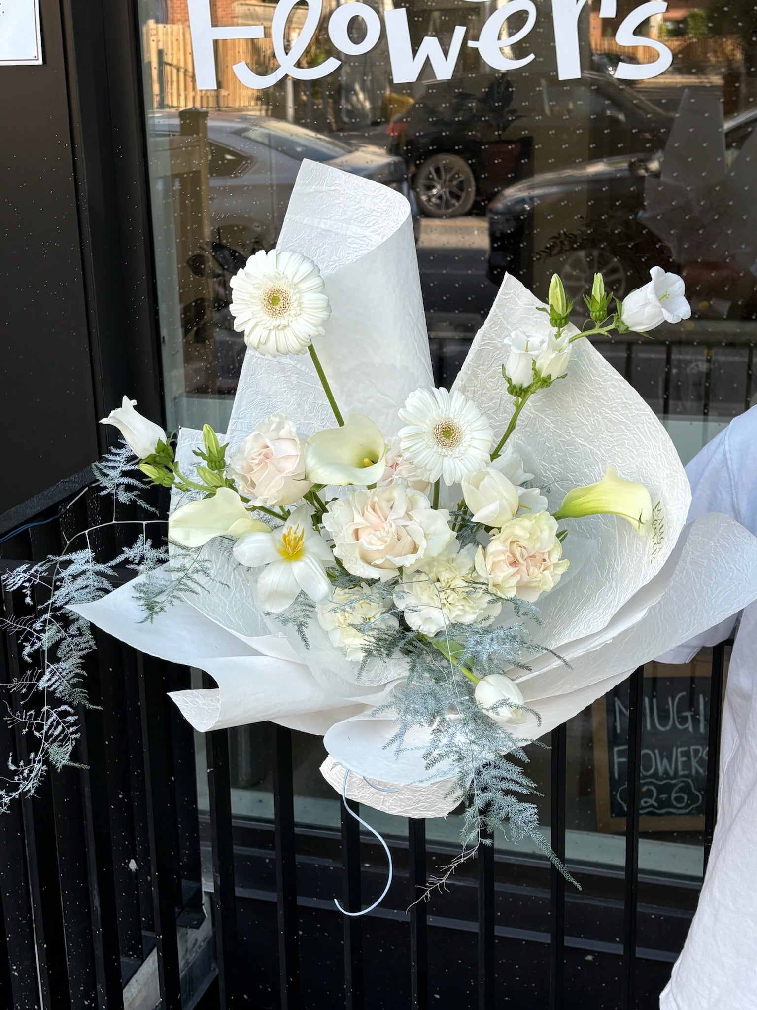 White bouquet featuring gerbera daisies, roses, calla lilies, and lisianthus wrapped in textured white paper, displayed outside Miugi Flowers shop.