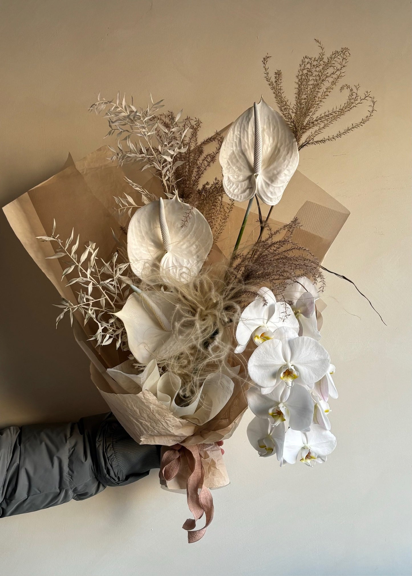 A hand holding a bouquet wrapped in brown kraft paper, featuring white anthuriums, dried foliage, and white orchids cascading at the front. The bouquet has a mix of textures with neutral tones and natural, minimalist wrapping.