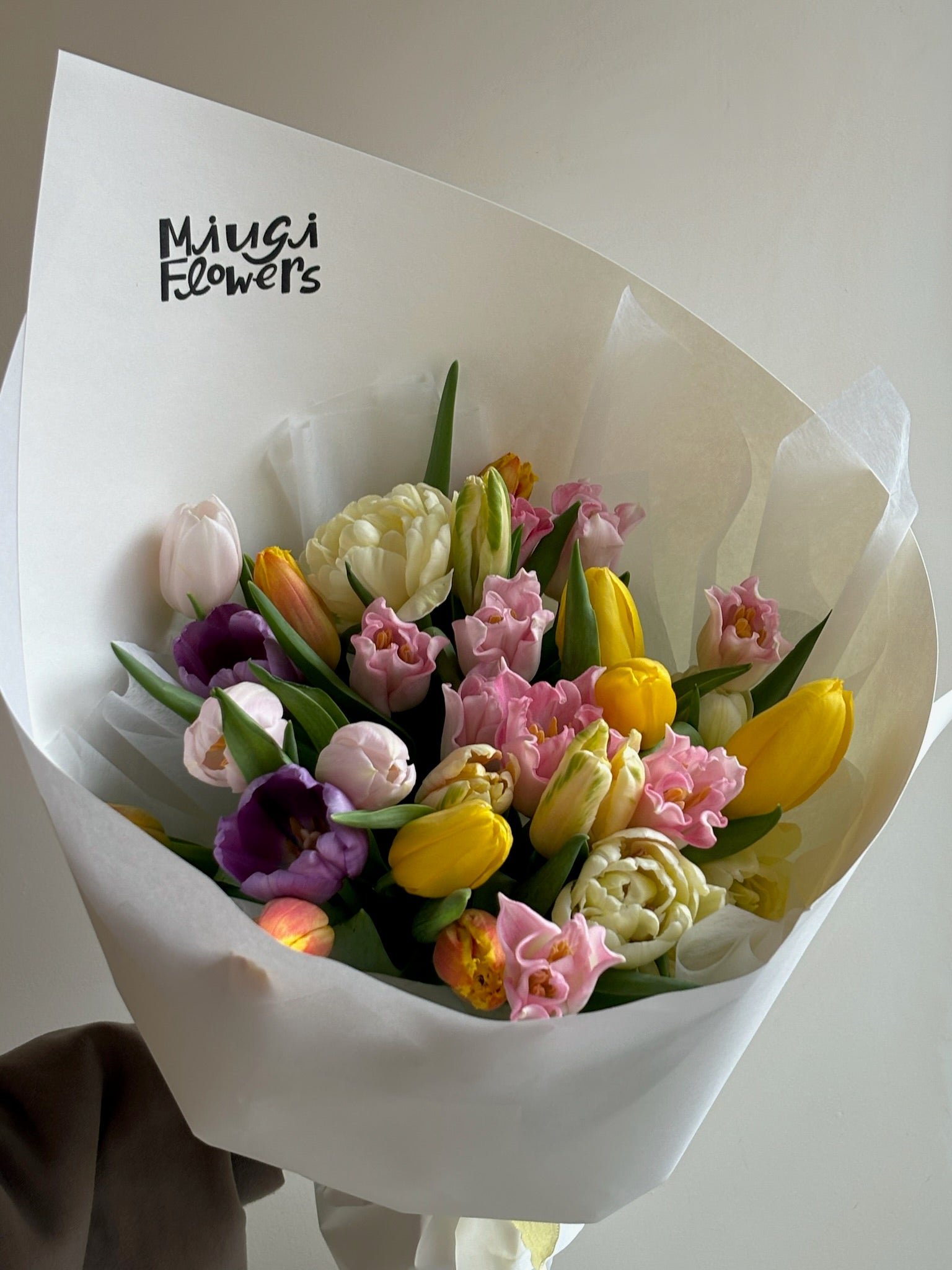 This image shows a floral bouquet wrapped in white paper with “Miugi Flowers” printed on the upper left corner of the wrapping. The bouquet contains tulips and other flowers in shades of yellow, pink, purple, and white. The flowers are arranged close together. The image background is plain.